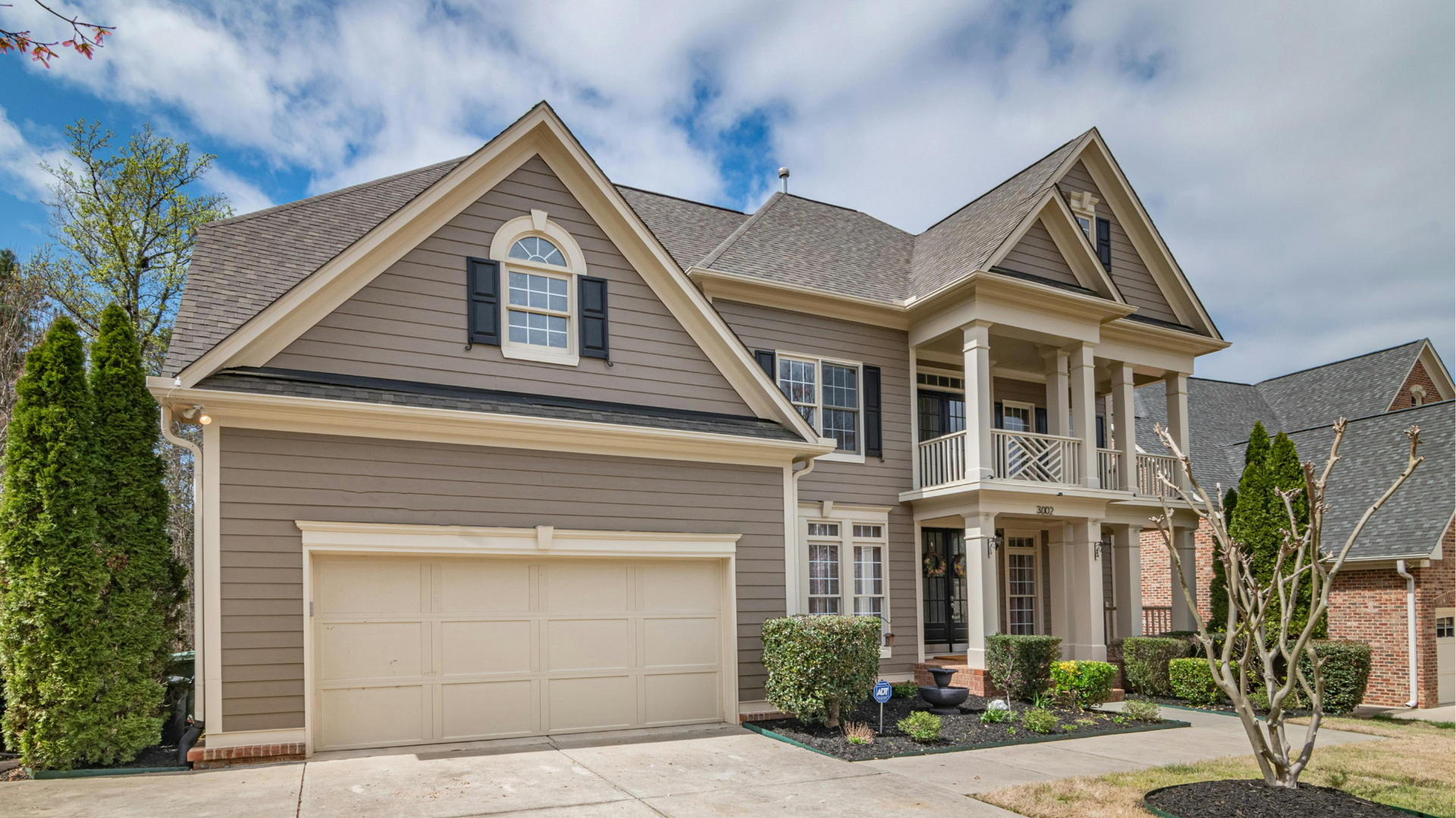 Two-story house with tan siding, columns, a garage, and landscaped yard under a cloudy sky.