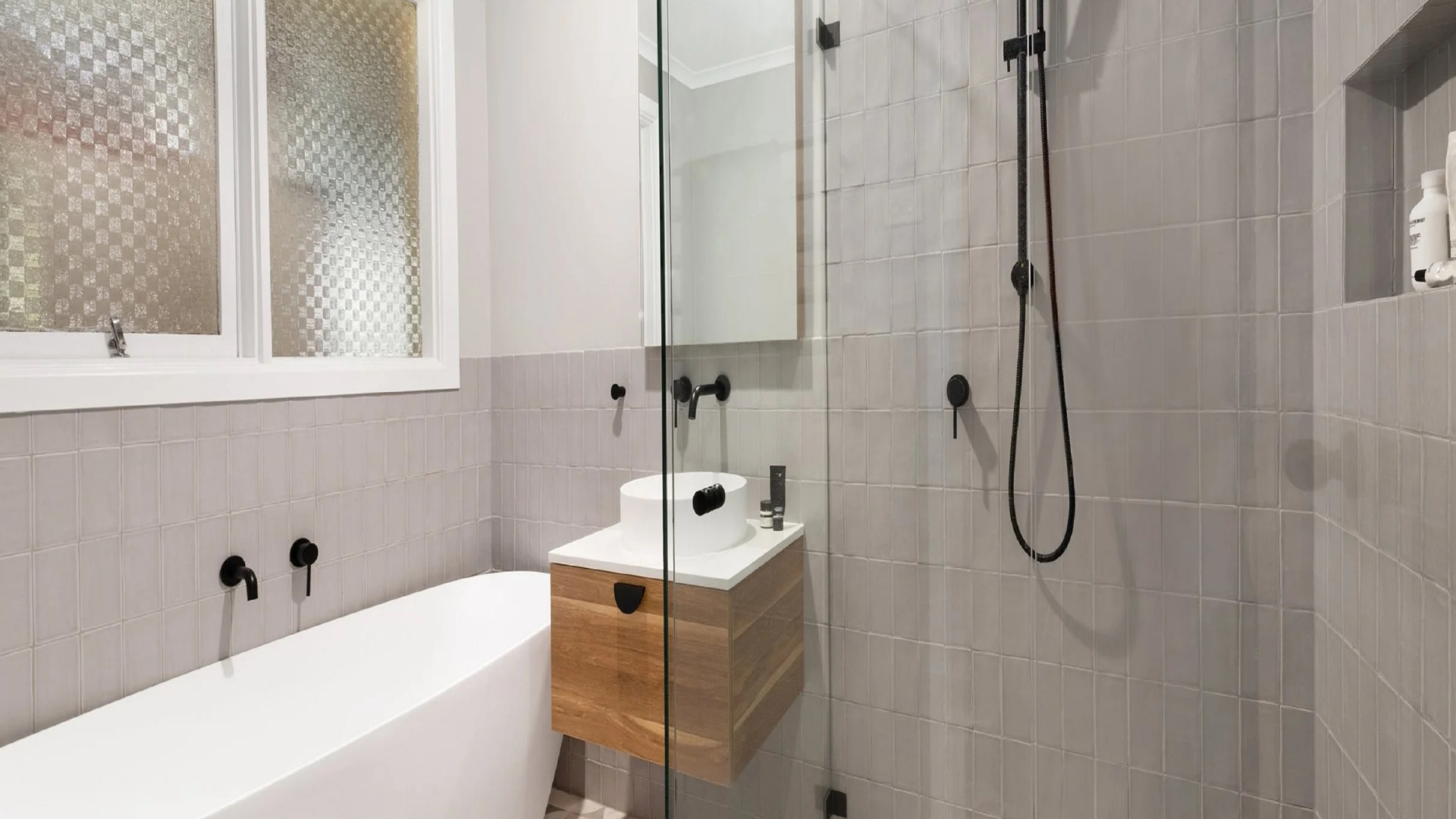 Modern bathroom with gray tiles, freestanding tub, glass shower, and wooden vanity.