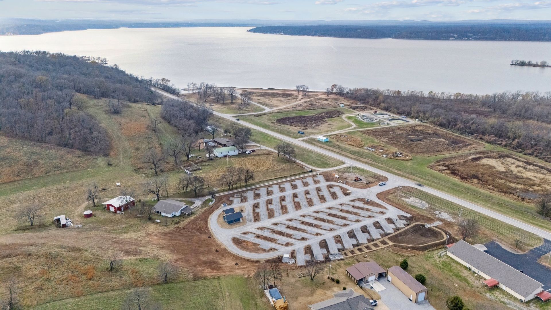 An aerial view of a lakeside RV park with rows of parking spots, nearby buildings, and surrounding wooded hills.