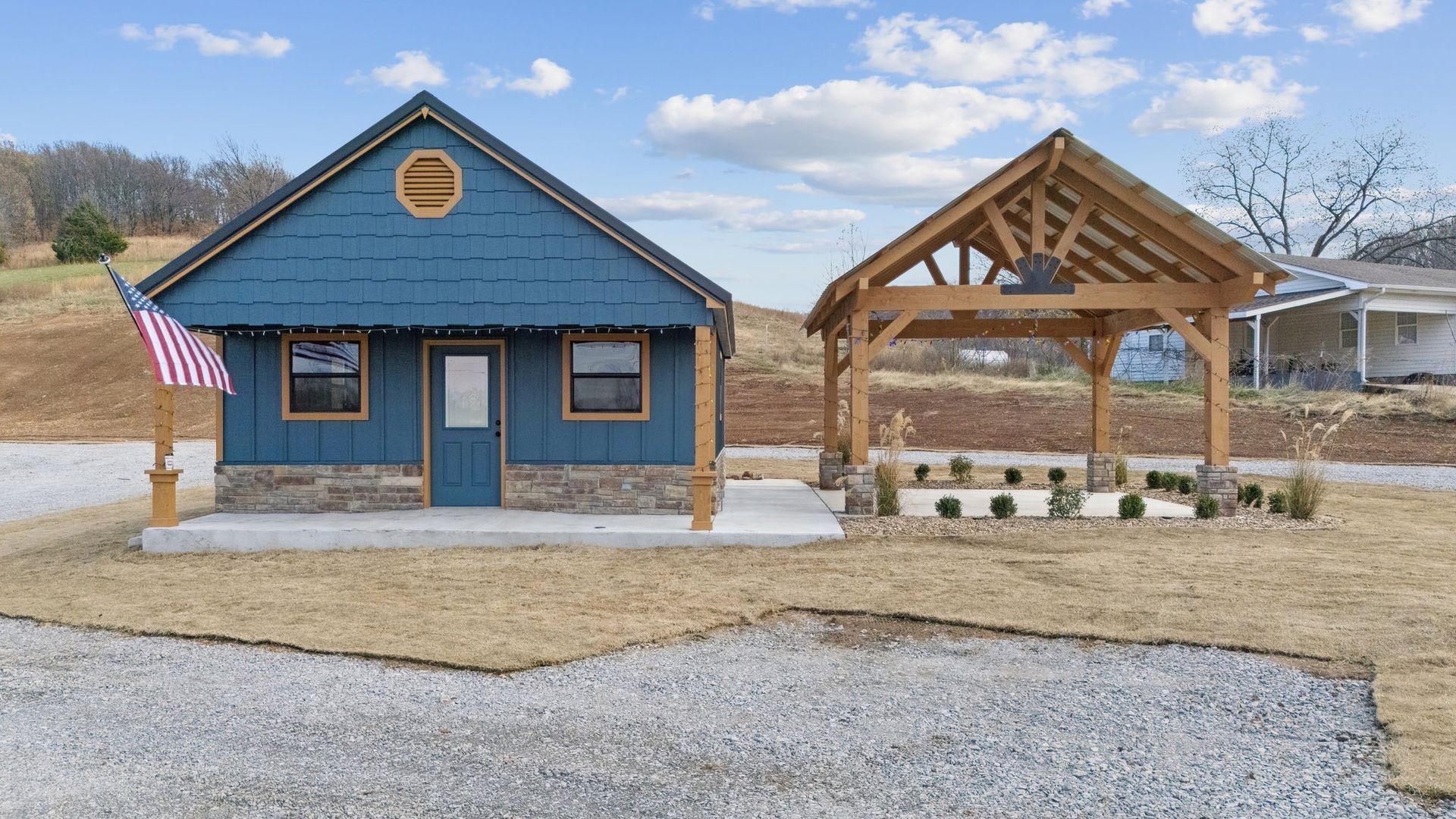 A blue house with a stone base and a matching timber-framed pavilion on a gravel lot under a sunny, cloudy sky.
