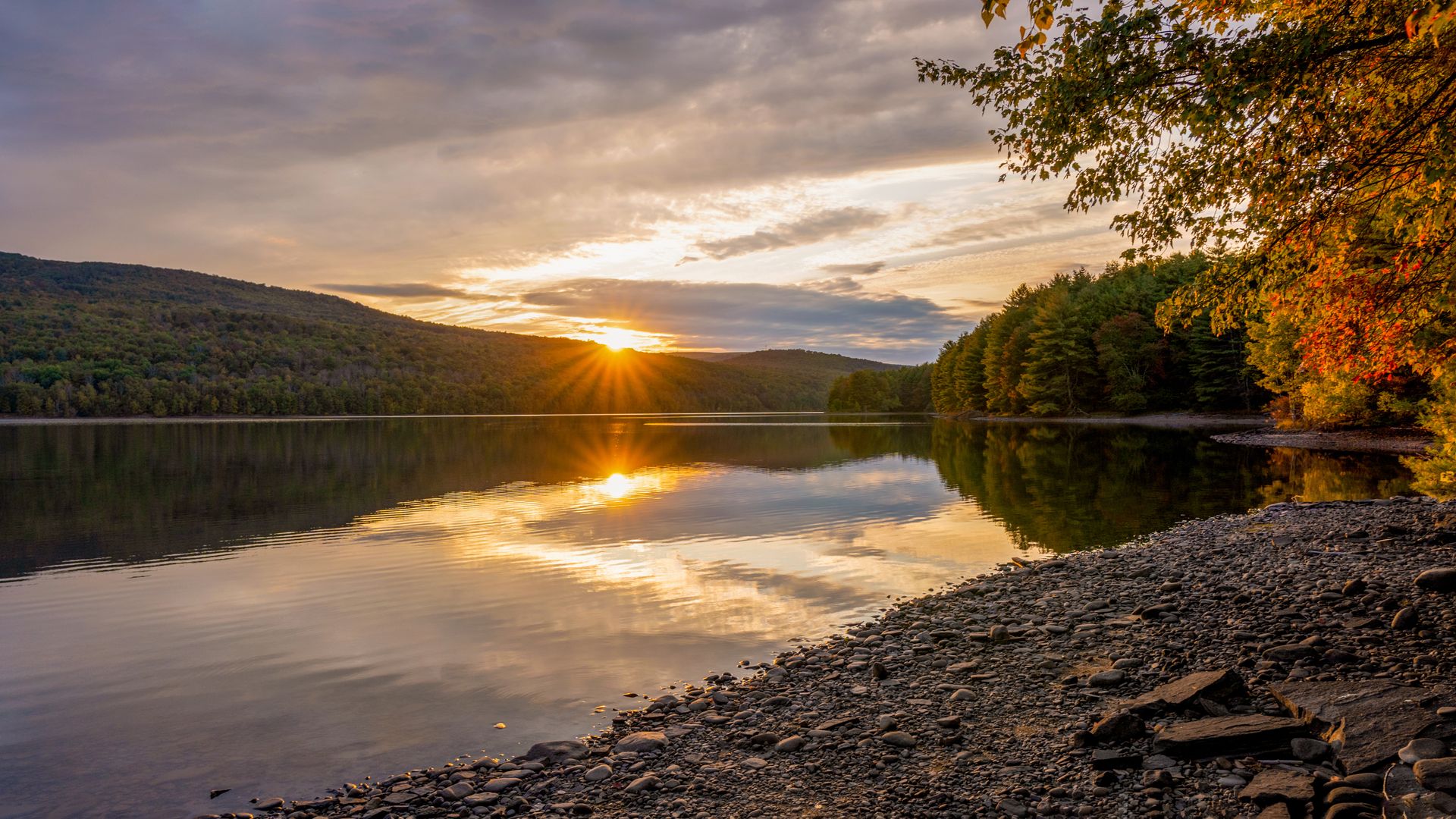 A tranquil lake reflects a golden sunset over tree-covered hills, with a rocky shoreline in the foreground.