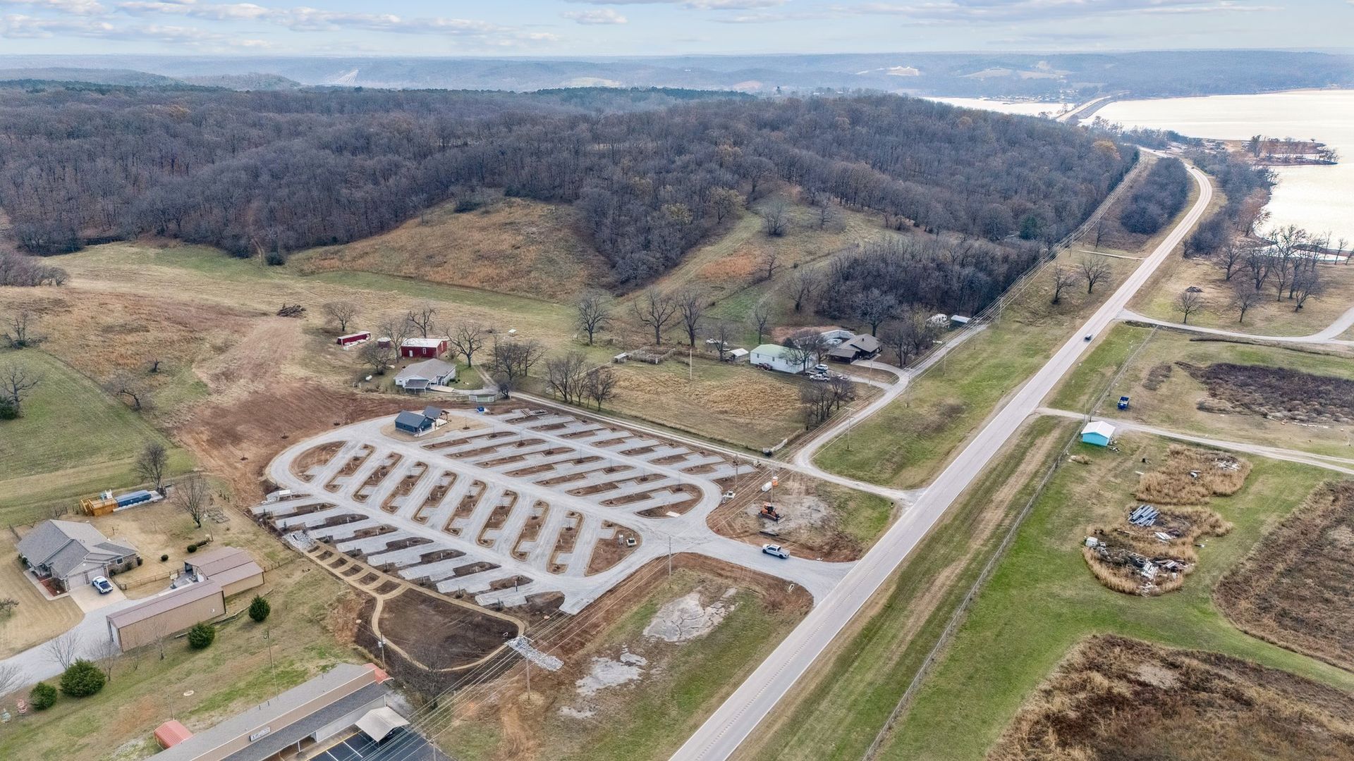 Aerial view of a paved boat ramp parking lot next to a road, field, and forested hillside near a body of water.