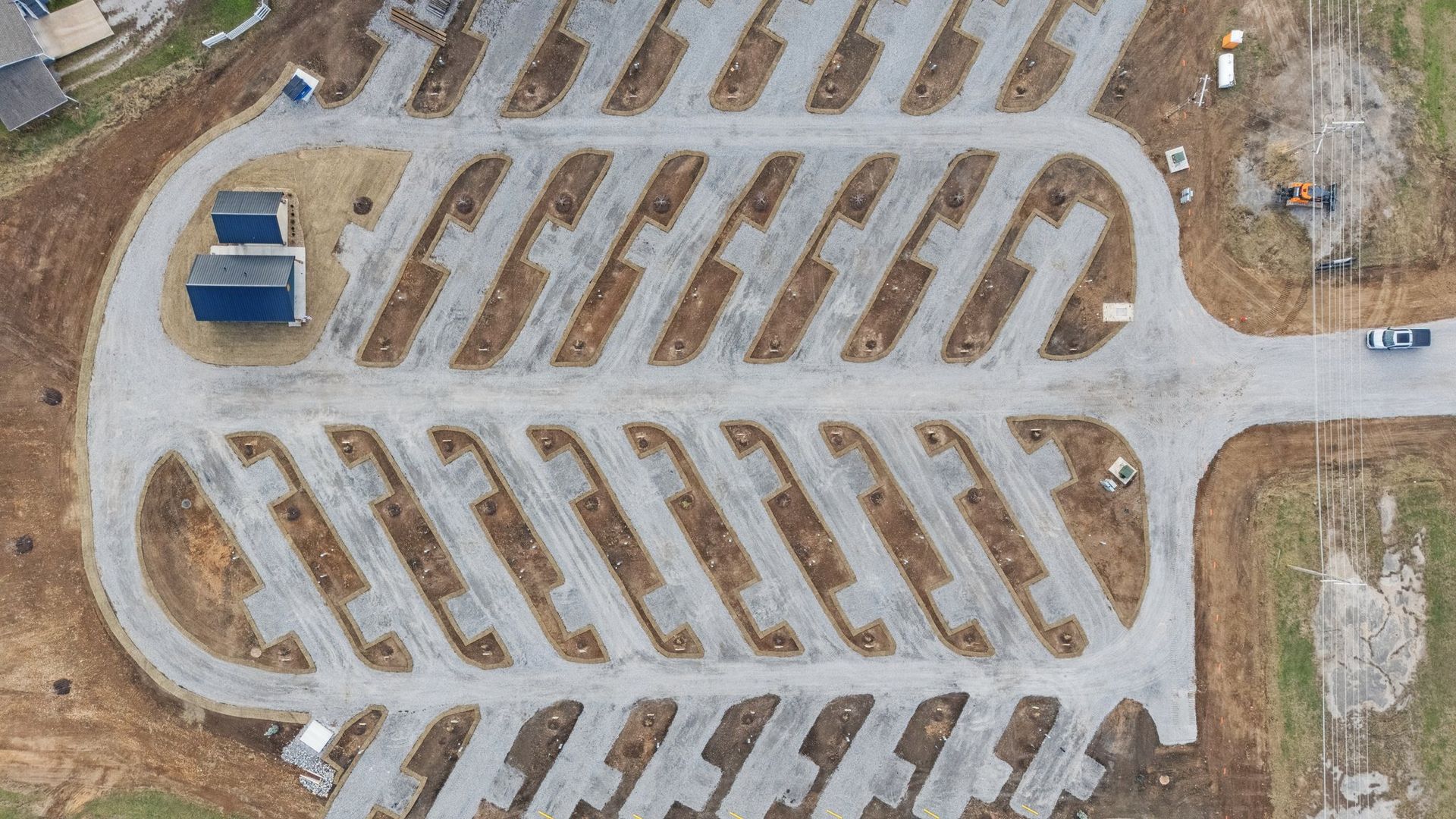 Aerial view of a gravel parking lot with diagonal parking spaces, surrounding a central roadway and two small buildings.