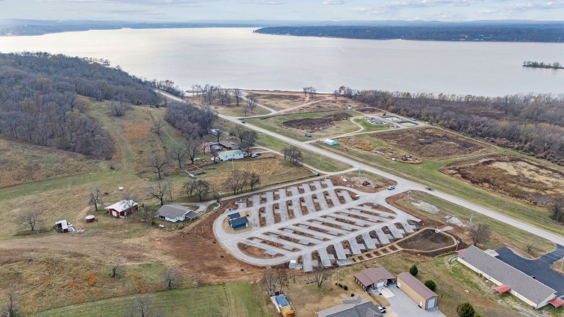 An aerial view shows an RV park with gravel spots and a central road, nestled in a grassy landscape beside a large lake.