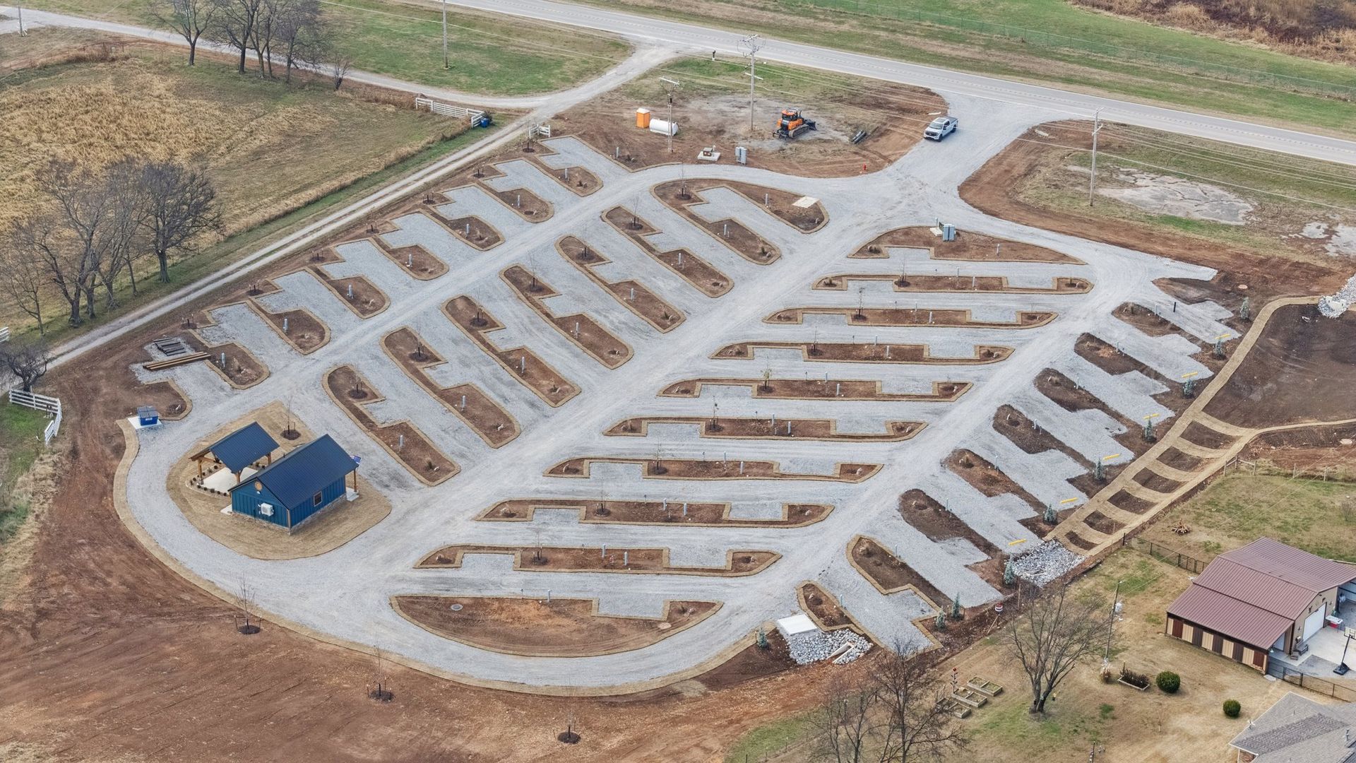 Aerial view of a gravel recreational vehicle park with marked camping sites, a central road, and a small service building.