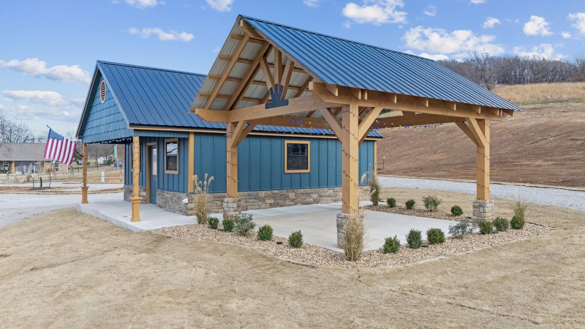 A blue metal-sided building with a stone base and an open-timber frame patio pavilion under a blue metal roof.