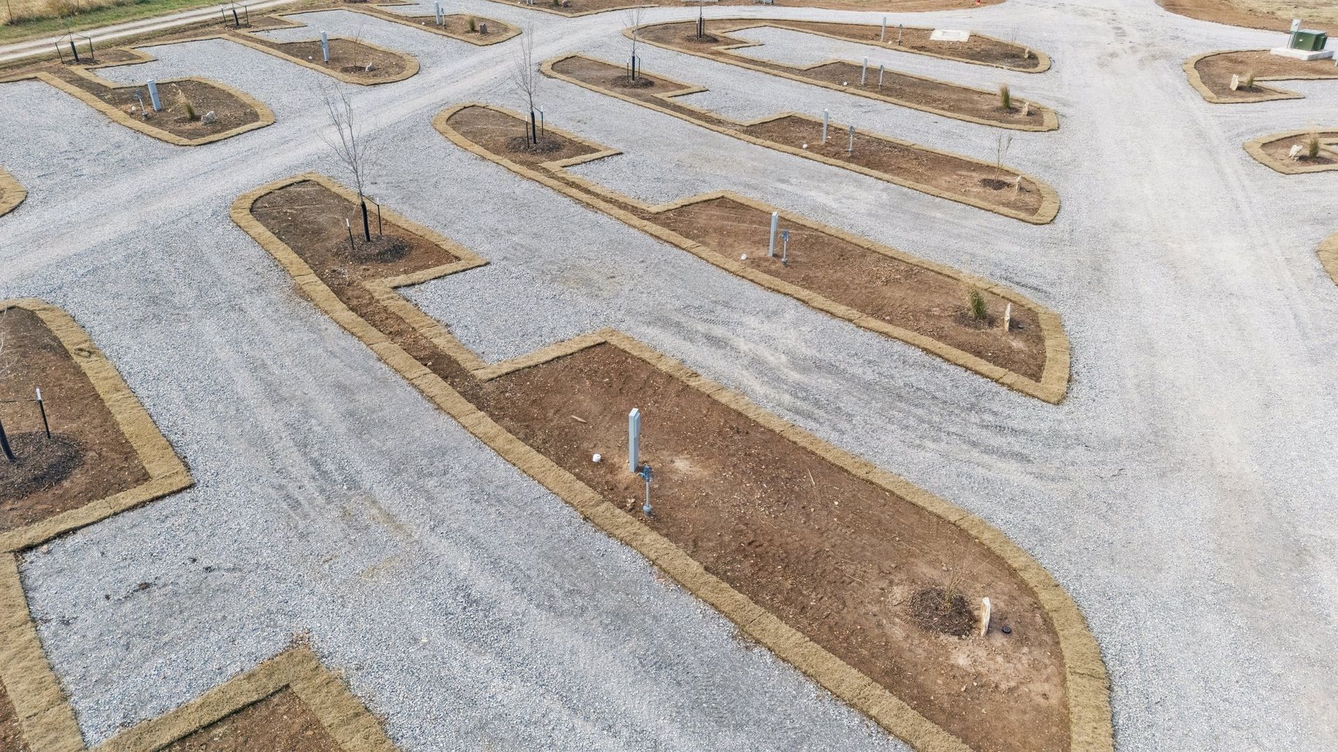 An aerial view of a graveled RV park with individual parking pads separated by dirt garden beds and small young trees.