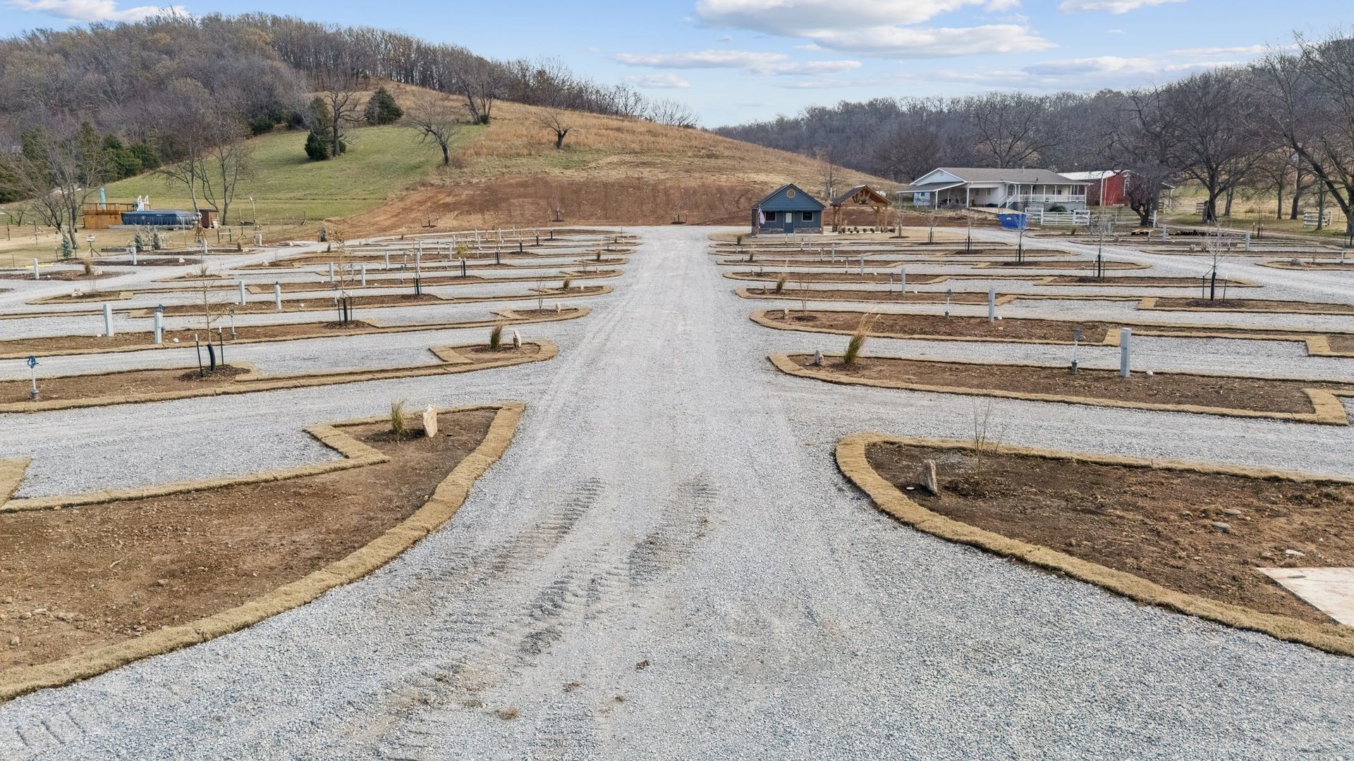 A gravel RV park or campground with multiple empty site pads, landscaping borders, and hills in the background.