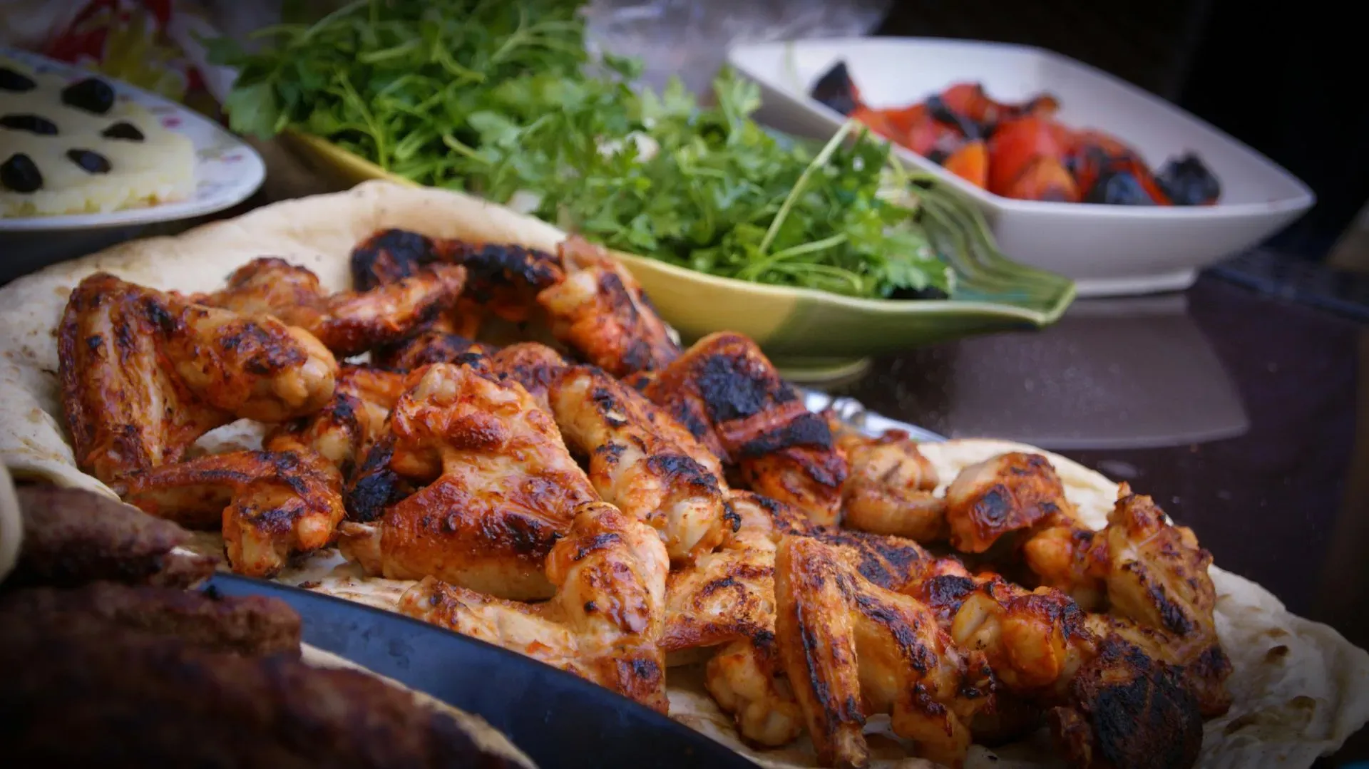 A platter of grilled chicken wings served on flatbread, with bowls of fresh greens and grilled tomatoes nearby.