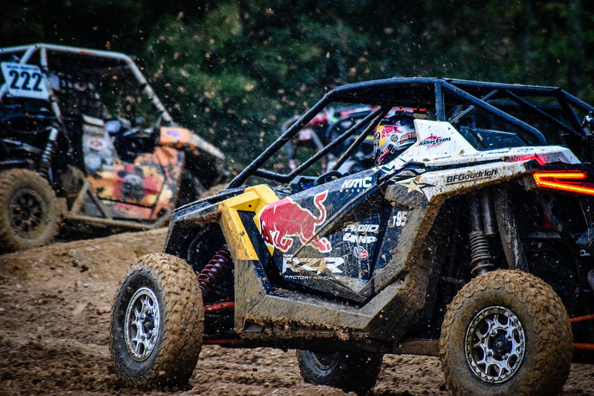 A side-angle view of two off-road race vehicles maneuvering through a muddy track, the lead car displaying a Red Bull logo.