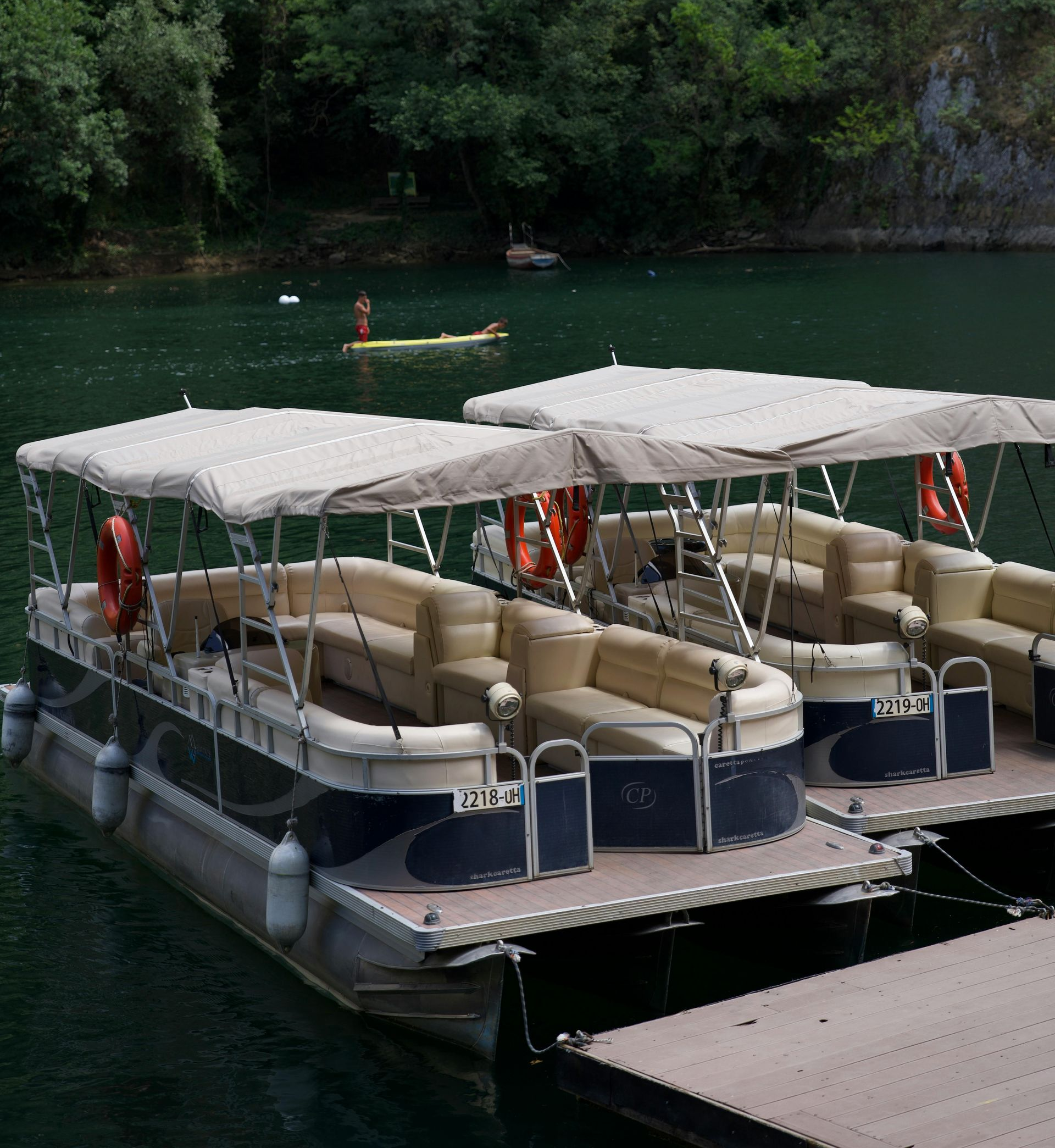 Two tan and navy pontoon boats moored at a wooden dock on a calm green river surrounded by trees.
