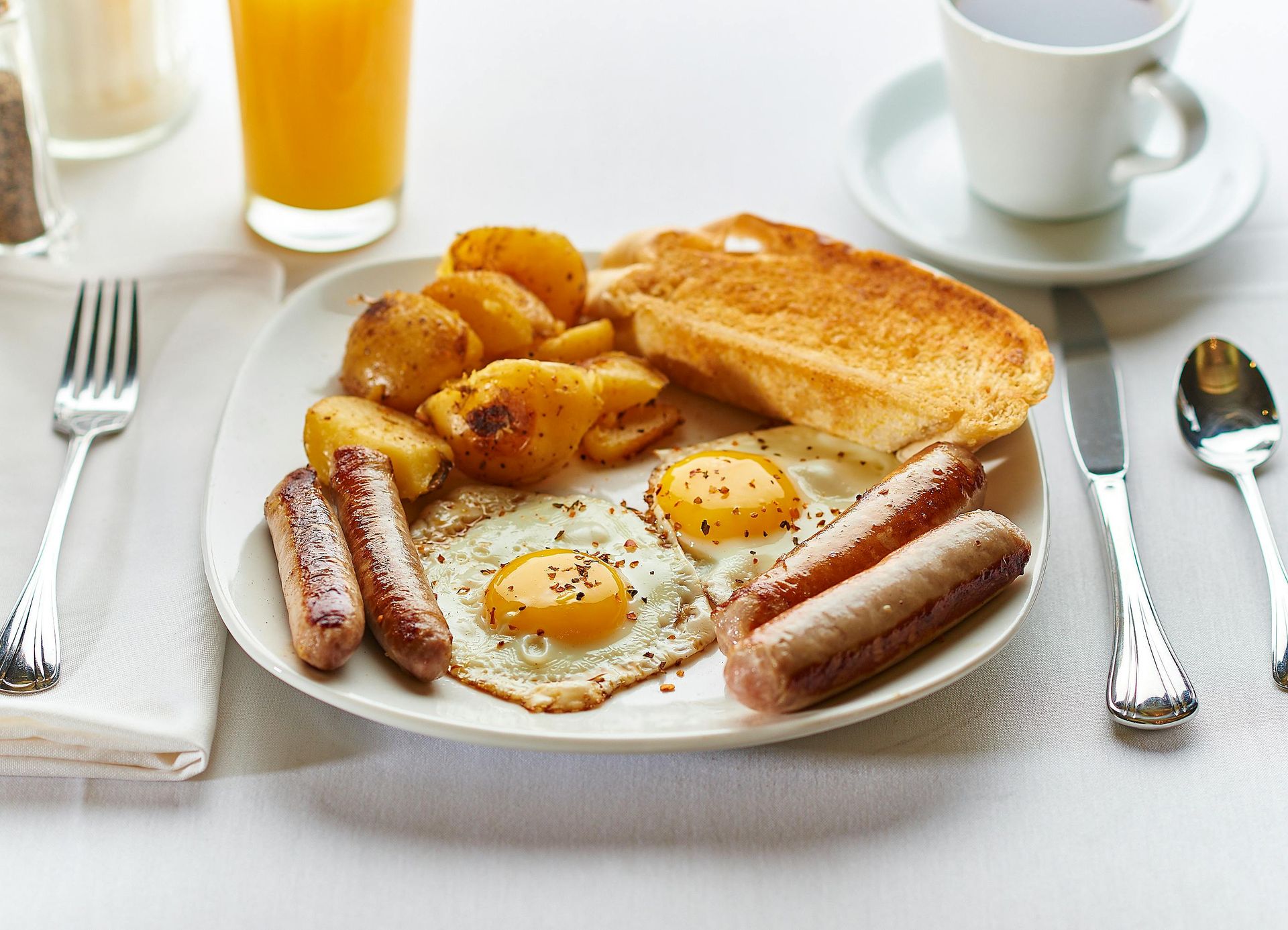 A breakfast plate with two eggs, four sausages, roasted potatoes, and toast, served with orange juice and coffee.