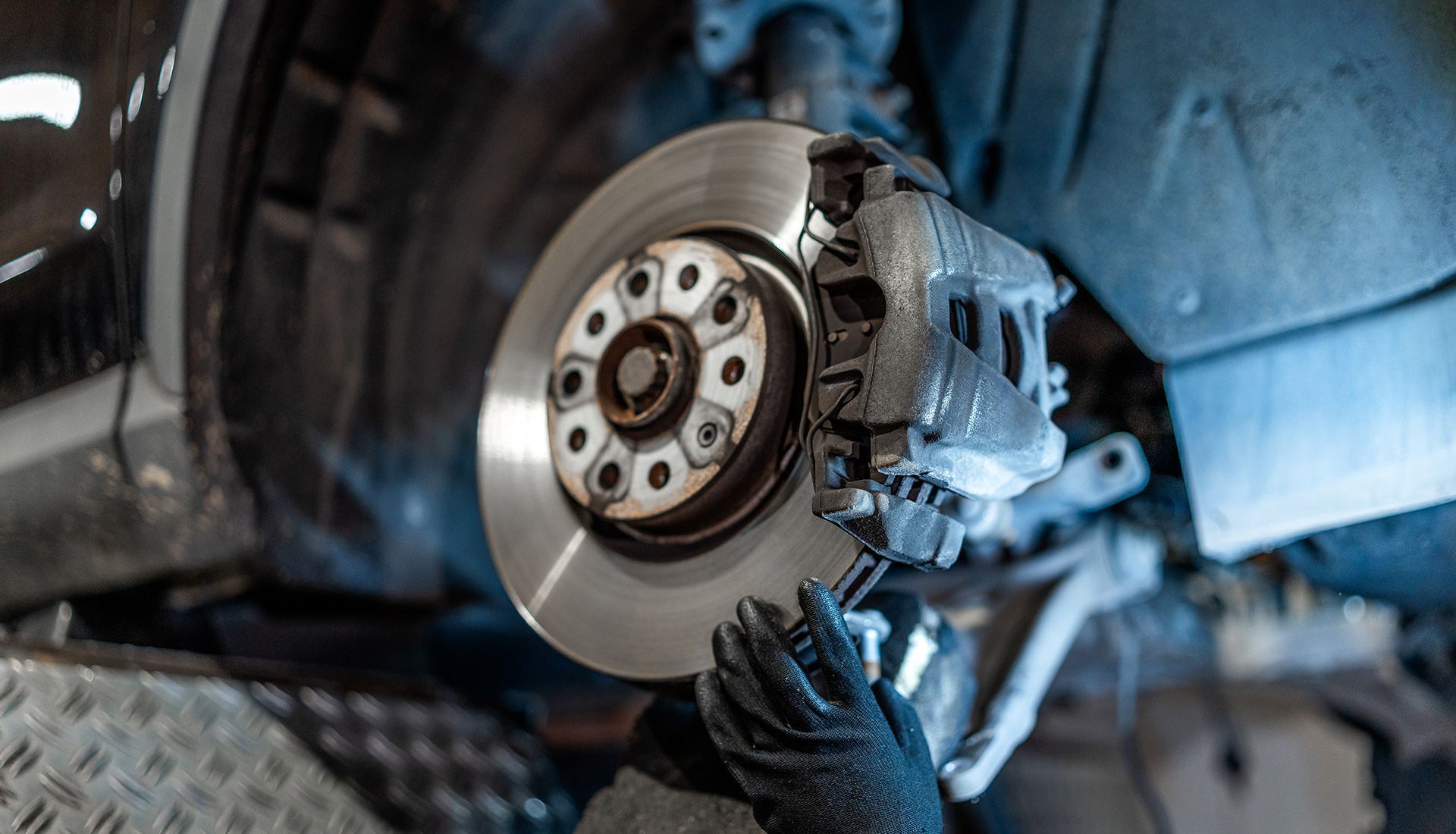 Mechanic working on a car's brake disc and caliper. Hands in gloves, wheel well visible.
