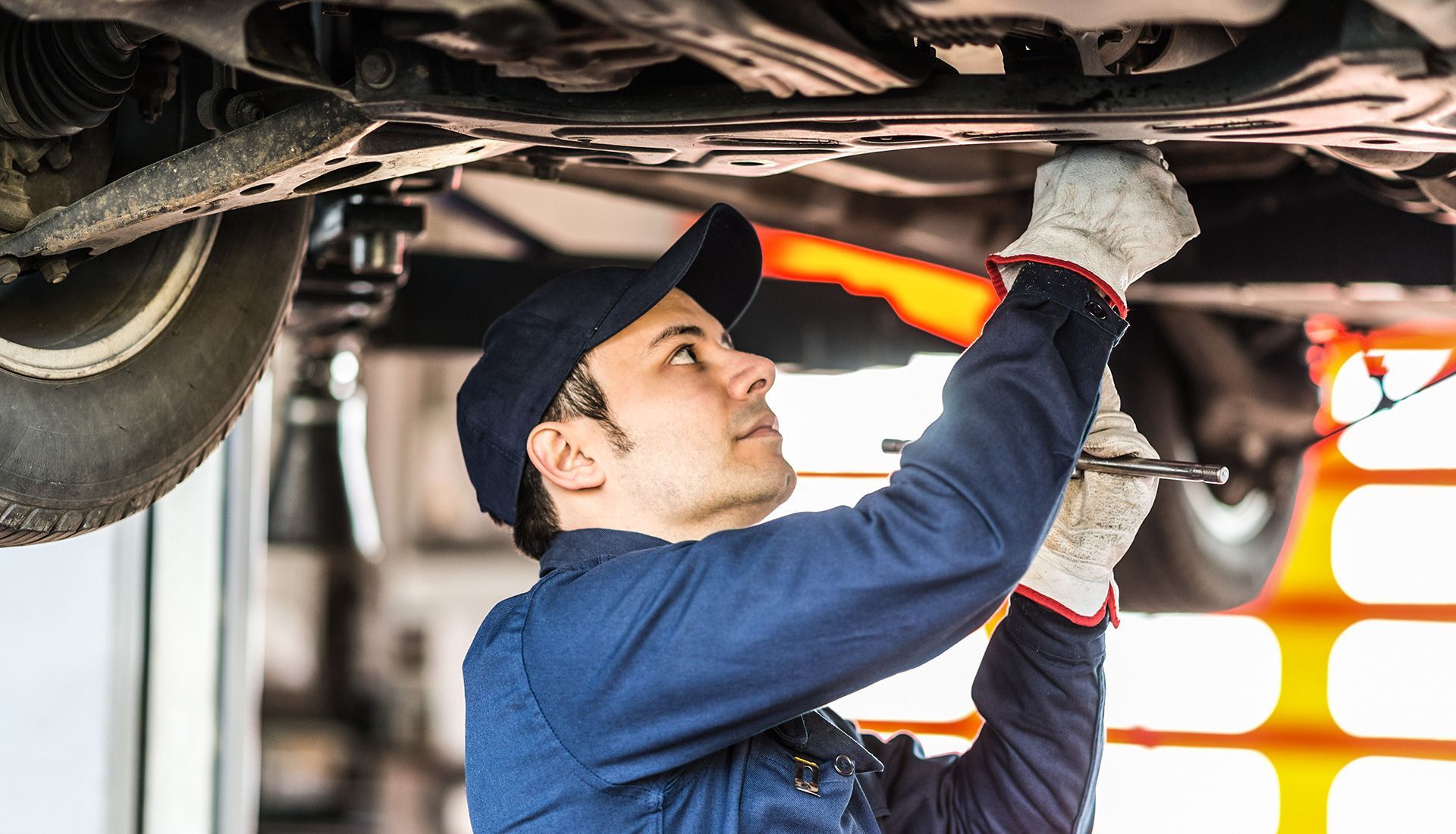 Mechanic working under a car, wearing a blue uniform, gloves, and a cap.