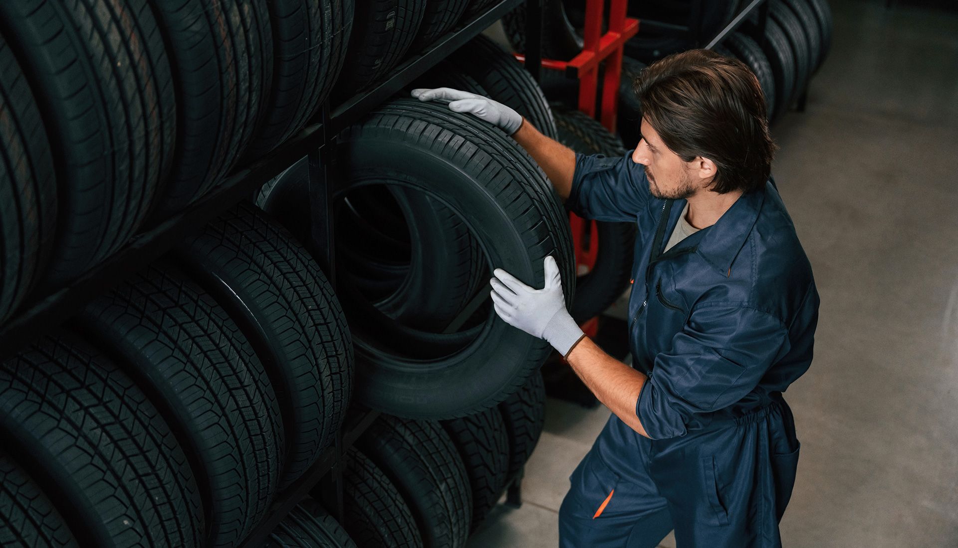 Mechanic in blue jumpsuit, wearing gloves, removes tire from a storage rack in a garage.
