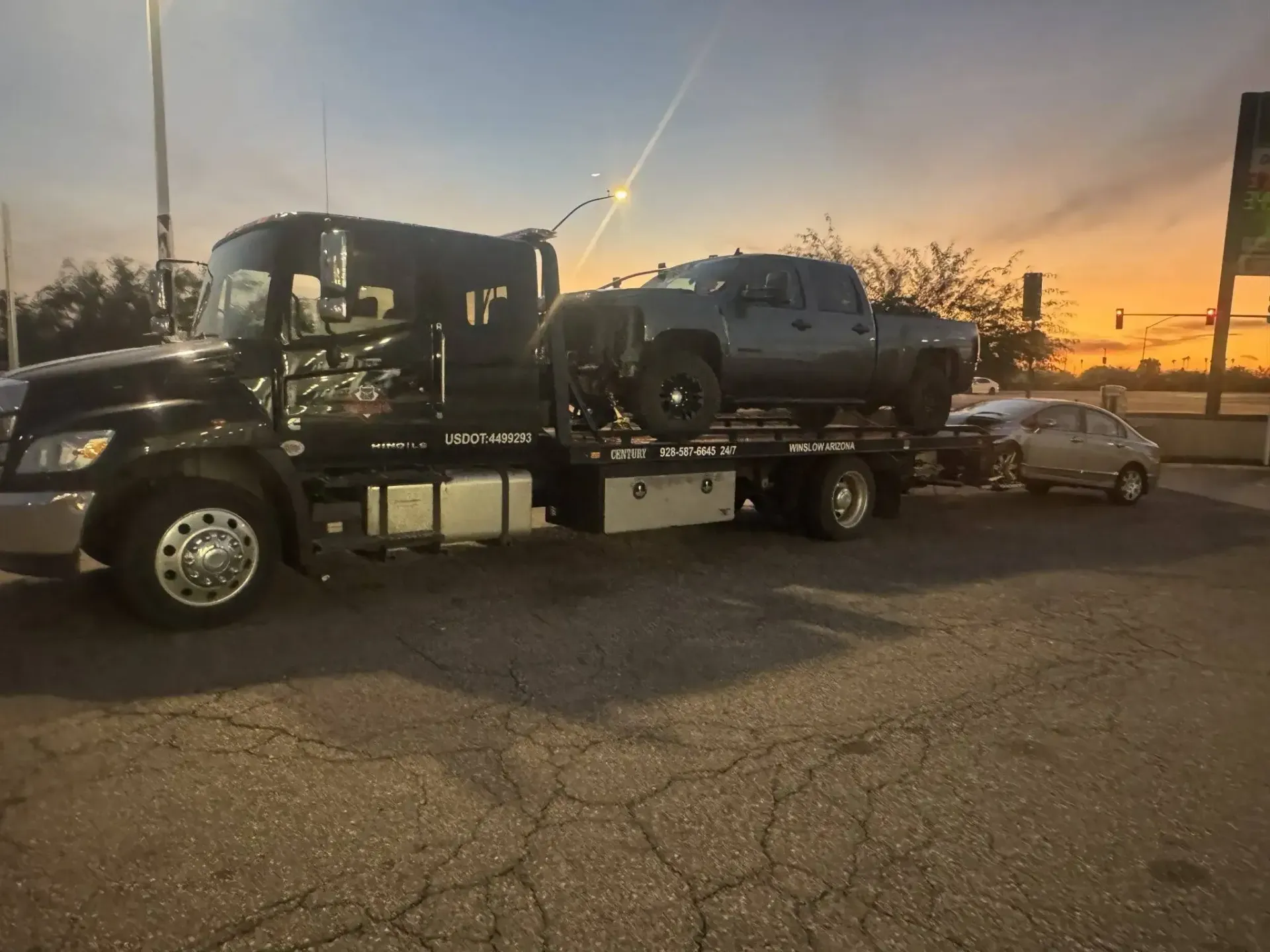 Tow truck carrying a black pickup at sunset, with a silver car behind it on a roadside.