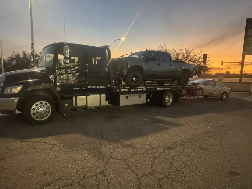 Tow truck carrying a black pickup at sunset, with a silver car behind it on a roadside.