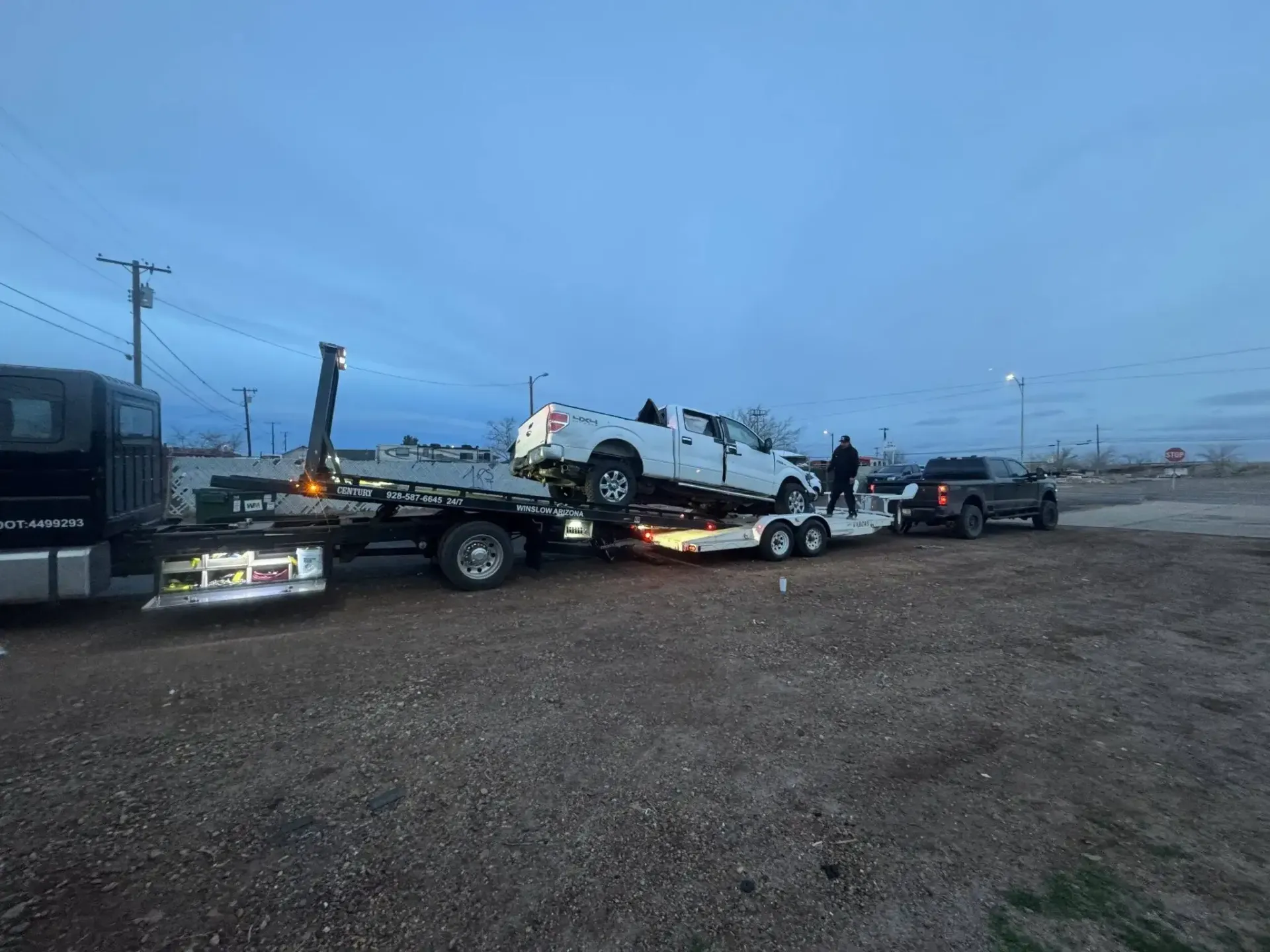 Tow truck hauling a white SUV on a gravel roadside at dusk, with another vehicle nearby
