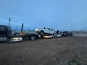 Tow truck hauling a white SUV on a gravel roadside at dusk, with another vehicle nearby