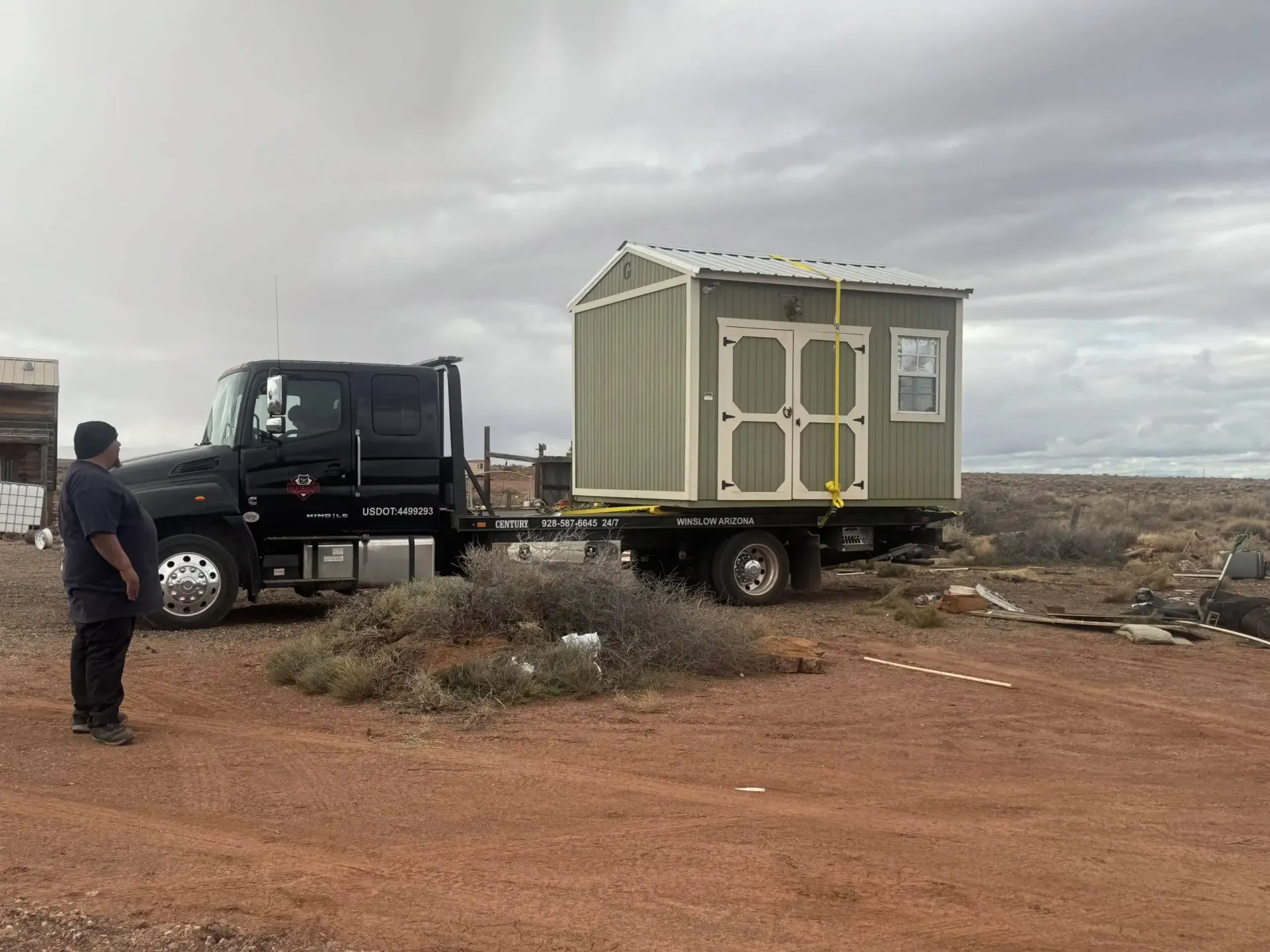 Black truck hauling a small off-road cabin on a dirt road under cloudy skies