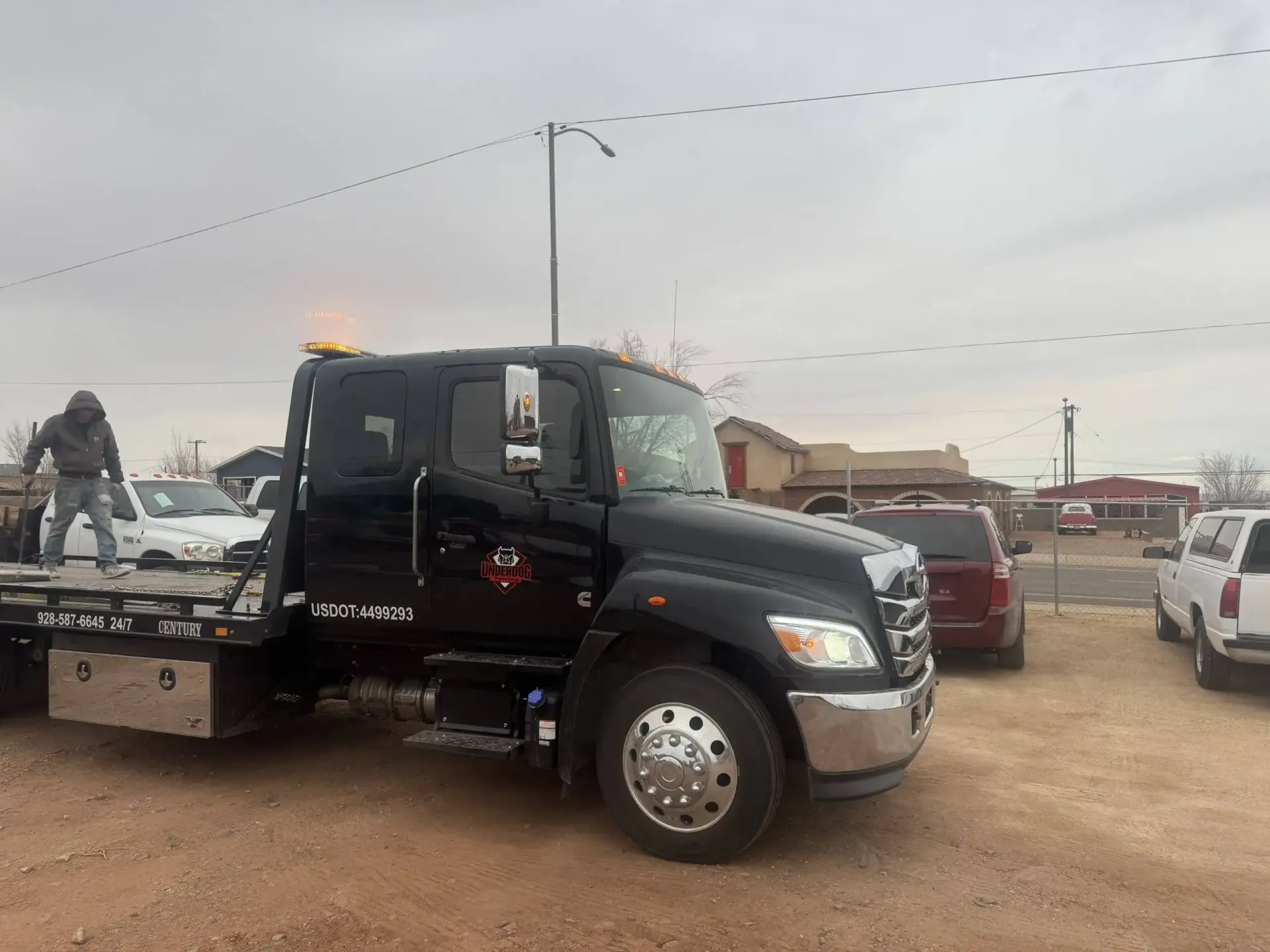 Black tow truck parked in a gravel lot beside other vehicles under an overcast sky