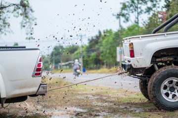 White pickup and SUV stuck in mud, connected by a tow strap on a wet dirt road