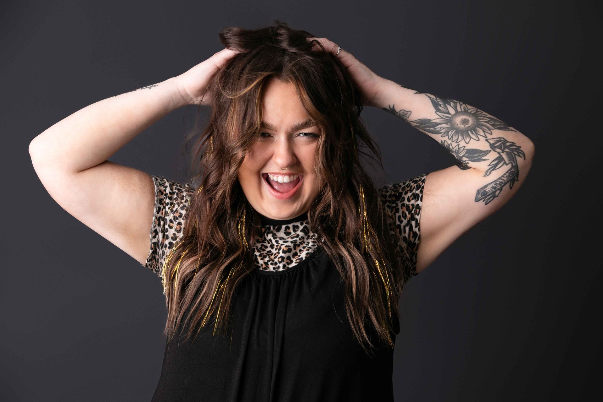 Person with long hair smiling, hands on head, wearing a black top against a dark background