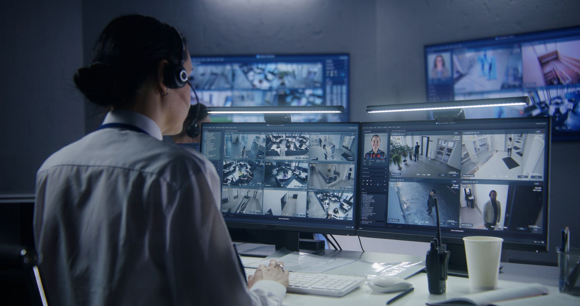 Person monitoring multiple security camera feeds at a desk in a dimly lit control room