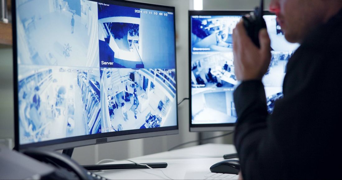 Person speaking into a radio in a security control room with multiple surveillance monitors.