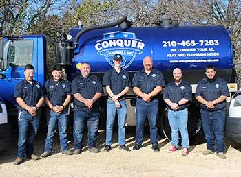Seven people stand in front of a blue truck with a logo for Conquer Plumbing and AC.