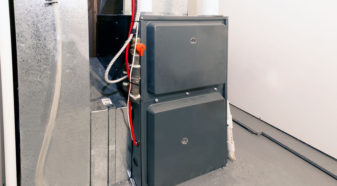 A furnace unit, gray metal, against a wall. Ductwork visible. Red and white piping.