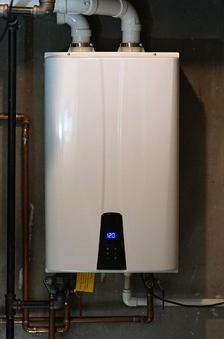 White tankless water heater mounted on a gray wall with copper and white PVC pipes.