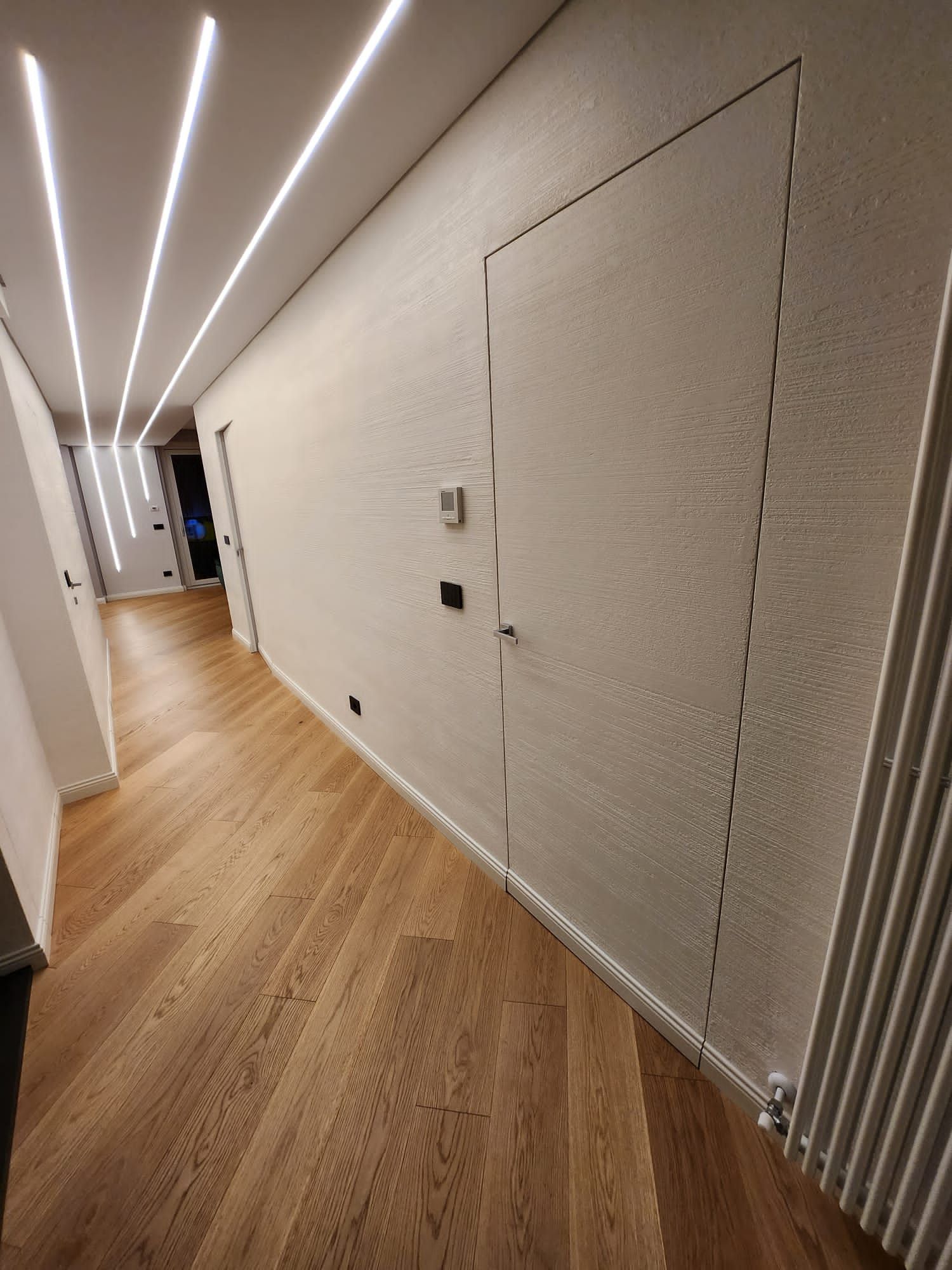 Corridoio stretto con pavimento in legno, pareti bianche testurizzate, illuminazione da incasso e una porta nascosta.