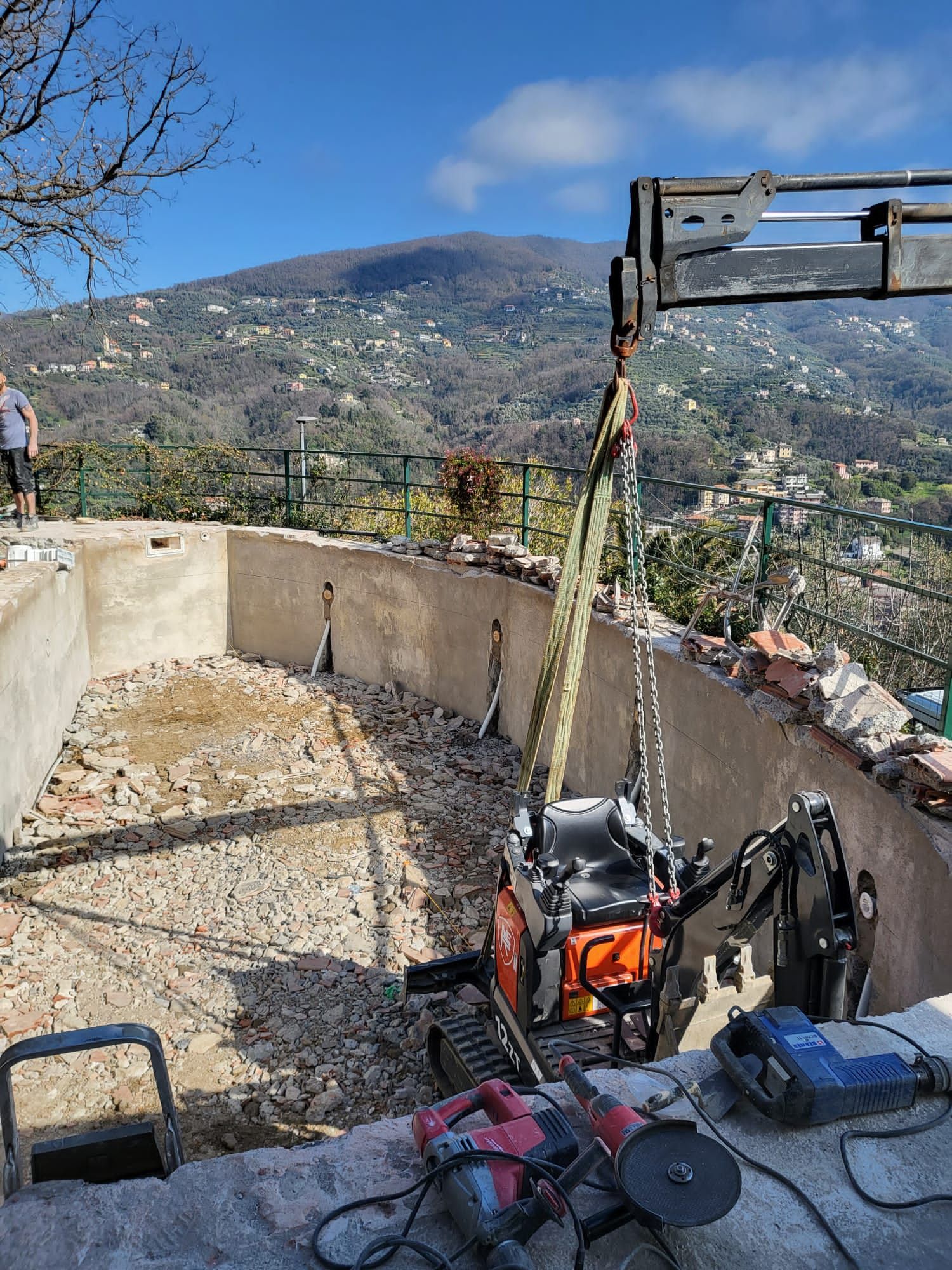 Miniescavatore sollevato da una piscina parzialmente demolita con vista panoramica sulle montagne.