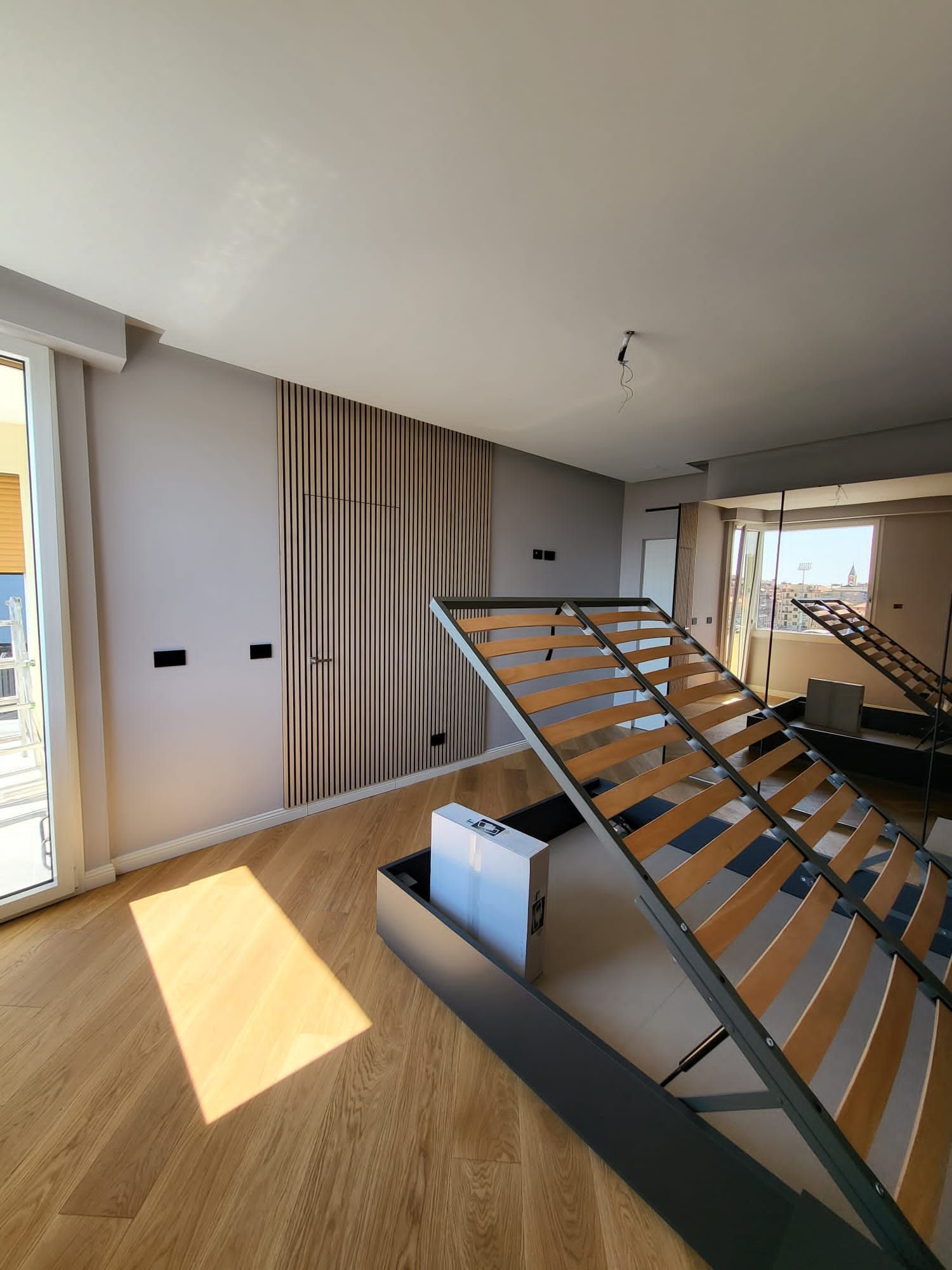 Interno della camera da letto con pavimento in legno, struttura del letto aperta, tende beige e luce solare.