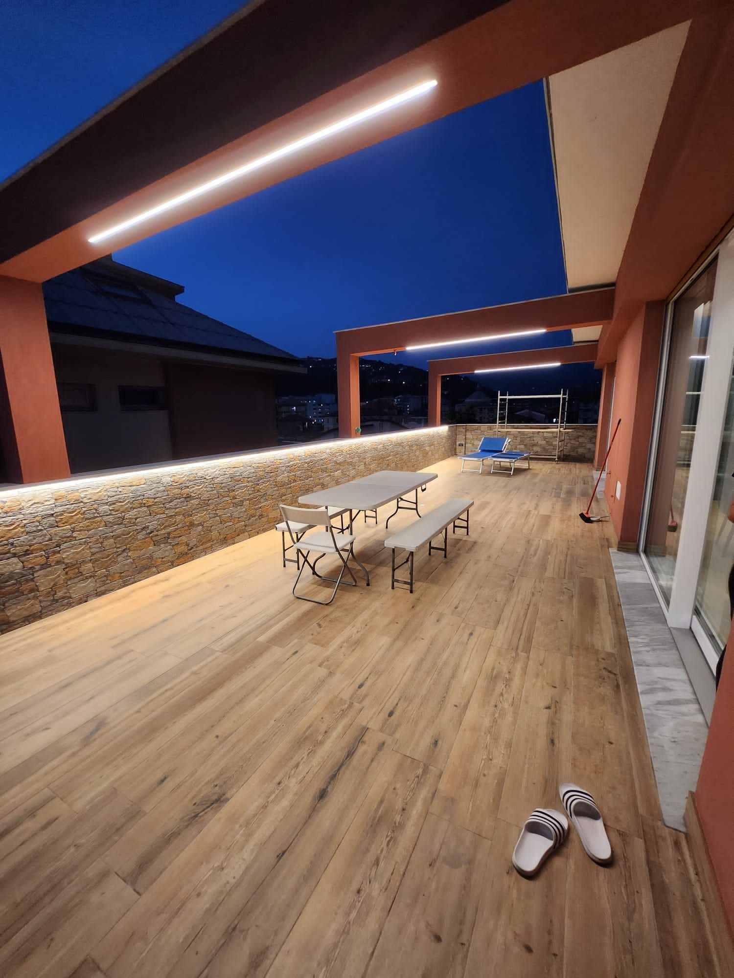Terrazza esterna in legno al tramonto, con tavoli, panche e illuminazione dall'alto.
