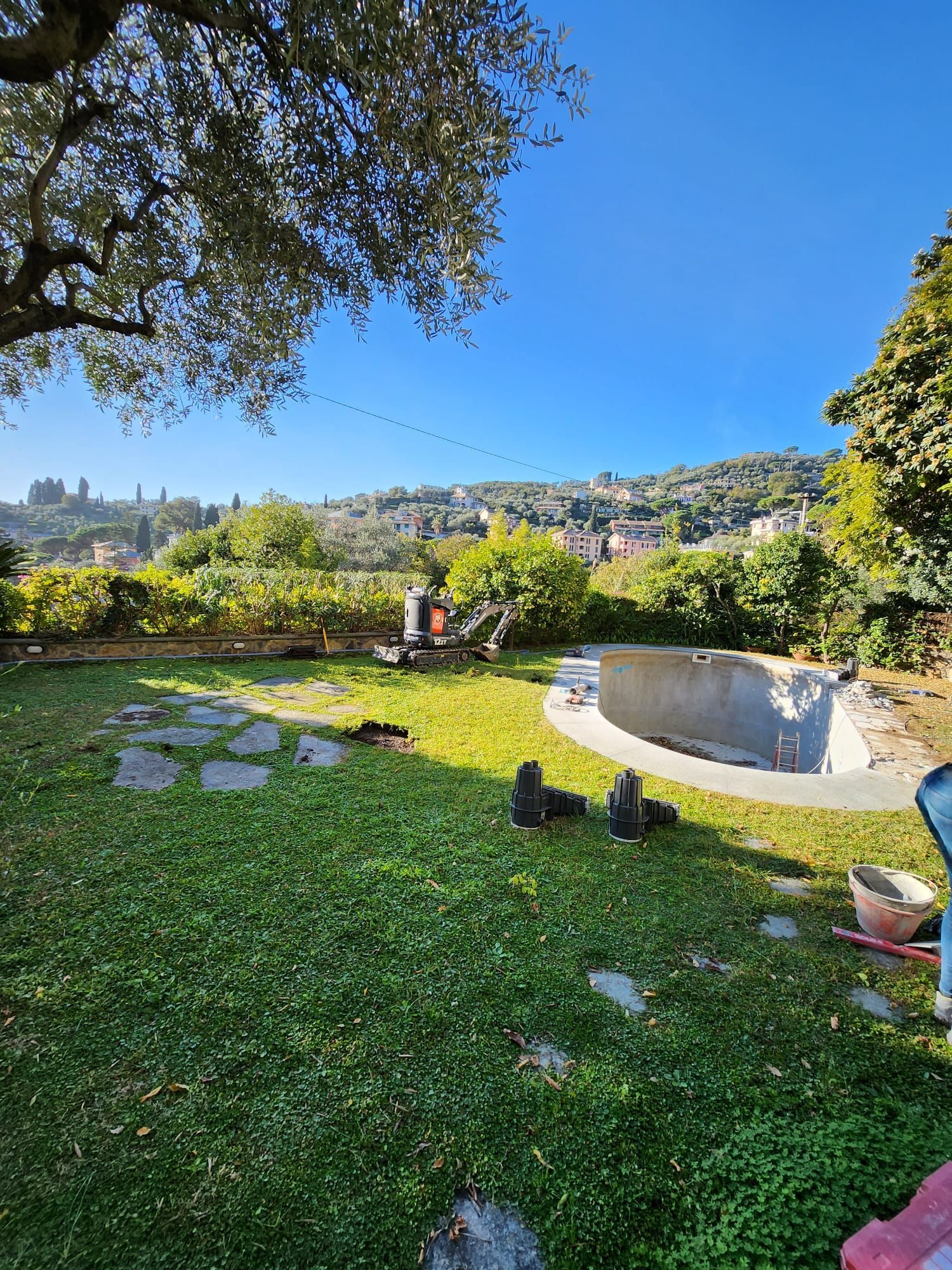 Giardino verdeggiante con sentiero in pietra, piscina parzialmente riempita e in lontananza una cittadina collinare sotto un cielo azzurro.