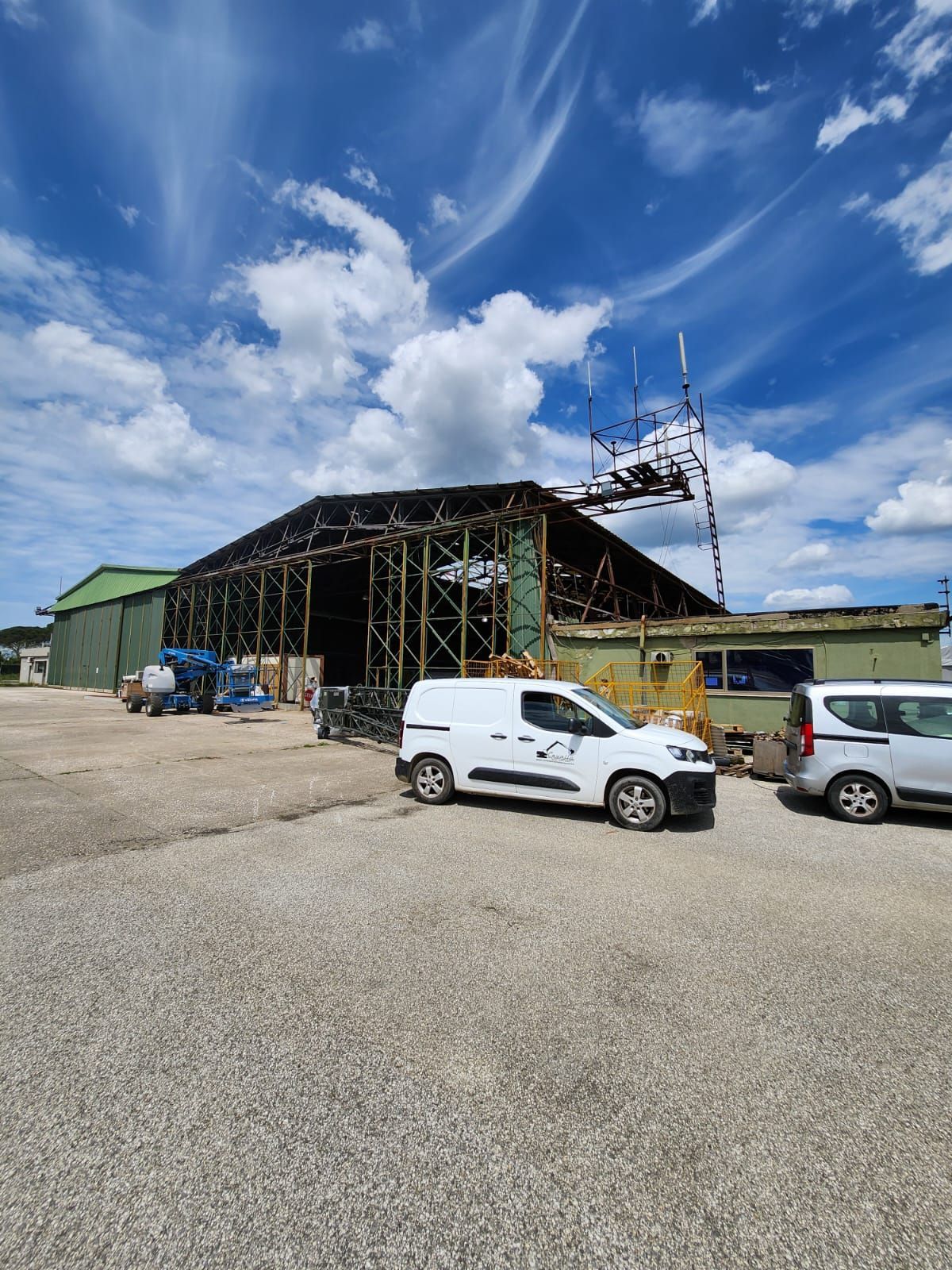 Hangar danneggiato con struttura scheletrica, furgoni parcheggiati, cielo nuvoloso.