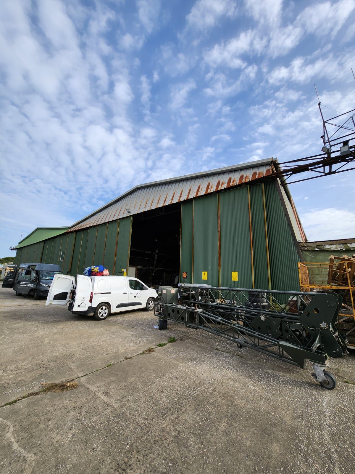 Hangar verde con porte aperte, veicoli parcheggiati all'esterno, cielo azzurro.