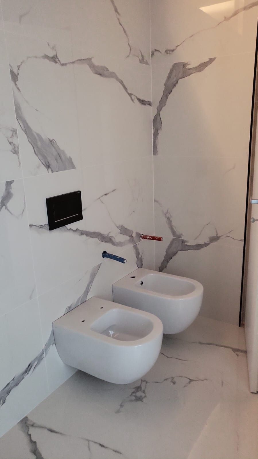 Due bidet bianchi su una parete piastrellata in marmo con venature grigie. Pannello di controllo nero, tubi rossi e blu.