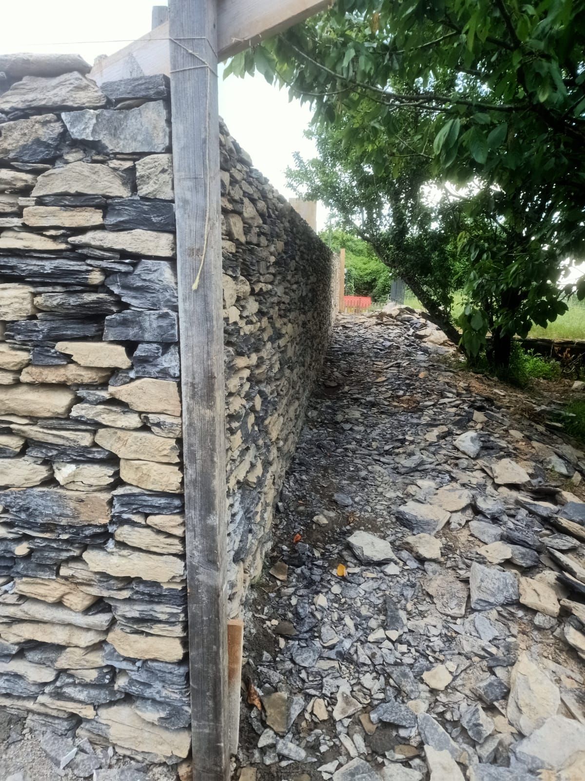 Muri in pietra incorniciano uno stretto sentiero, con un palo di legno e vegetazione sporgente.