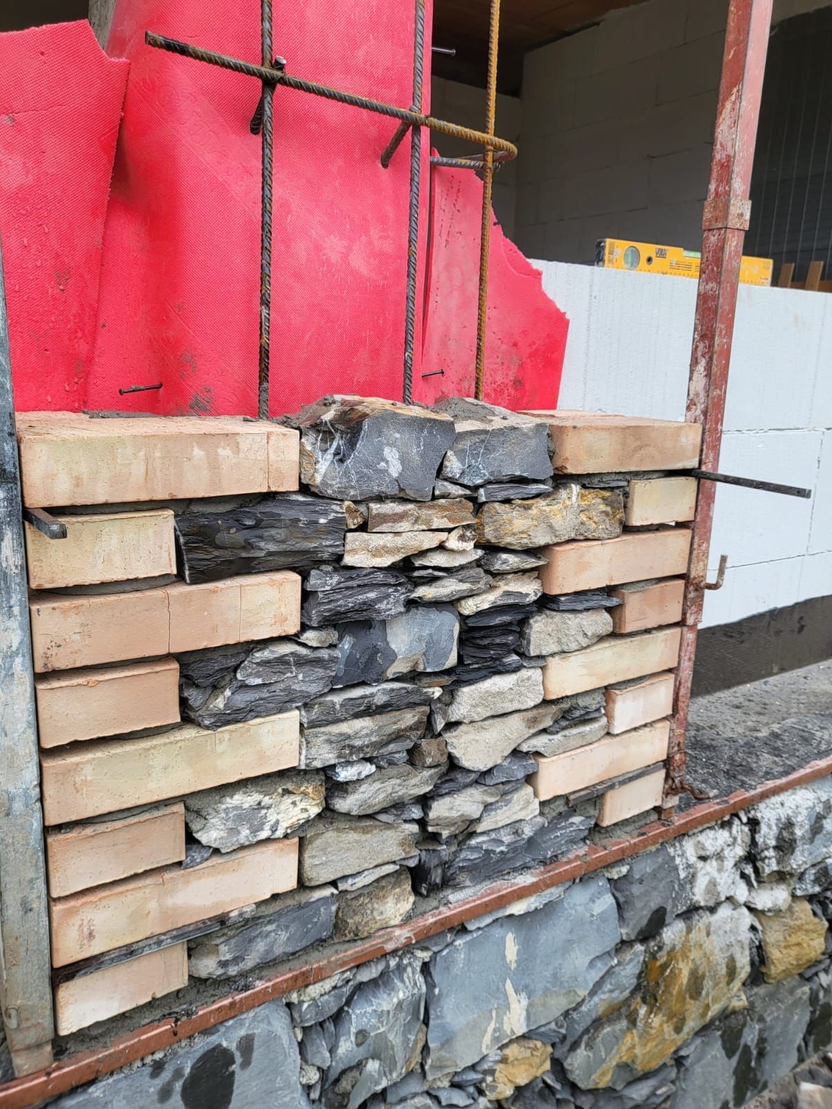 Muro in pietra e mattoni in costruzione con supporto in tondini di ferro e isolamento rosso.