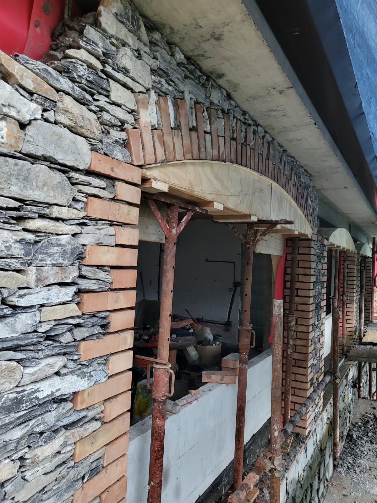 Facciata di un edificio in pietra e mattoni in costruzione con aperture ad arco; sostenuta da impalcature.