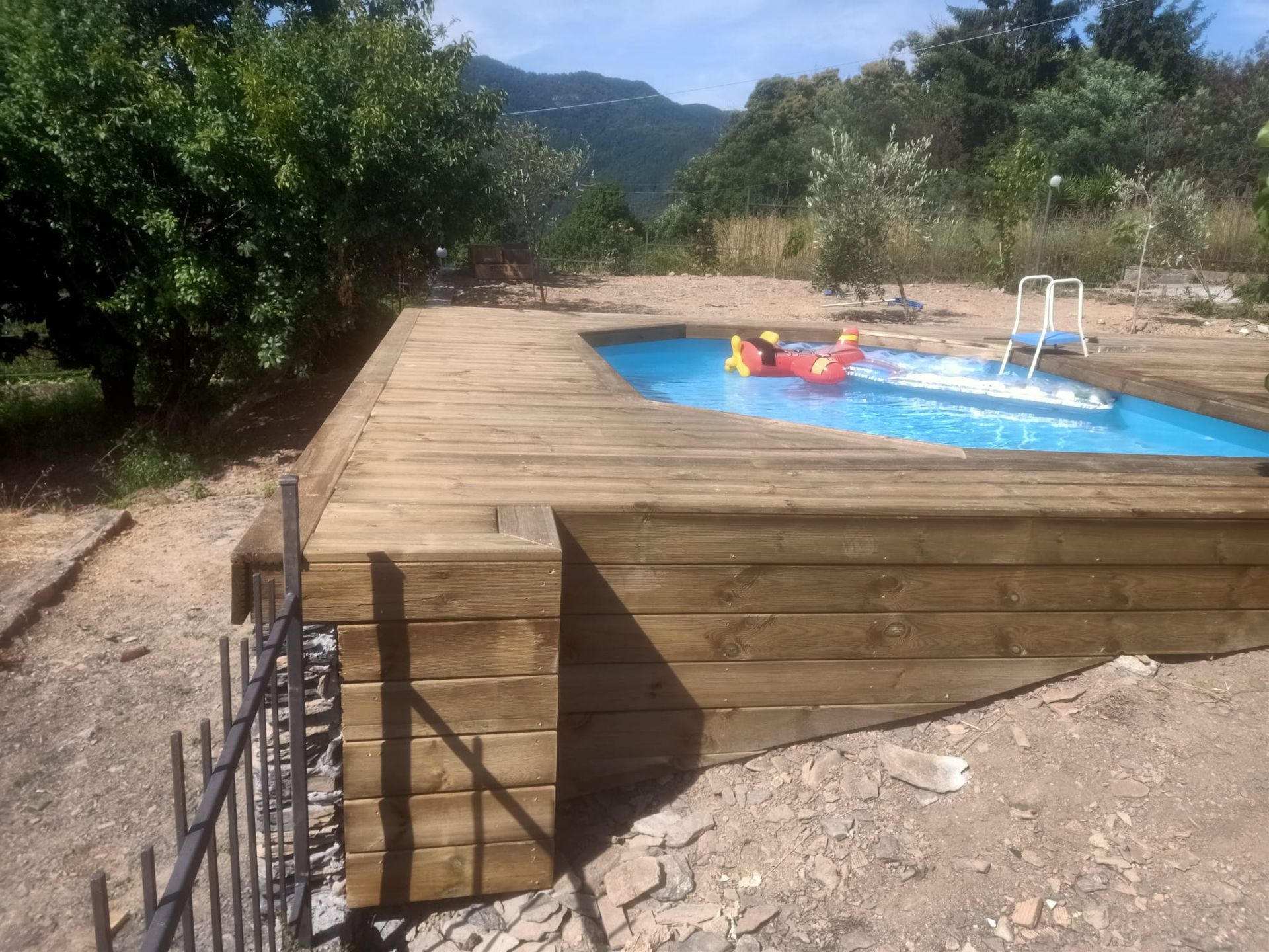 Terrazza in legno che circonda una piscina blu; montagne e vegetazione sullo sfondo, giornata di sole.