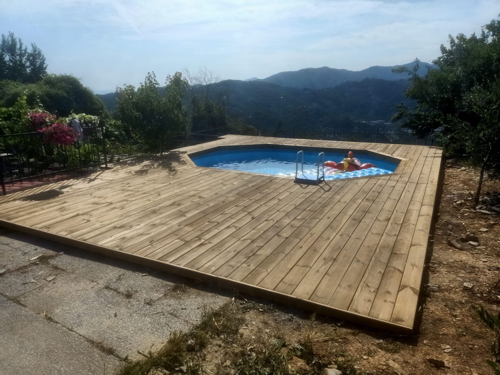 Un ponte di legno circonda una piscina blu con una persona su un galleggiante, con vista sulle montagne in una giornata di sole.