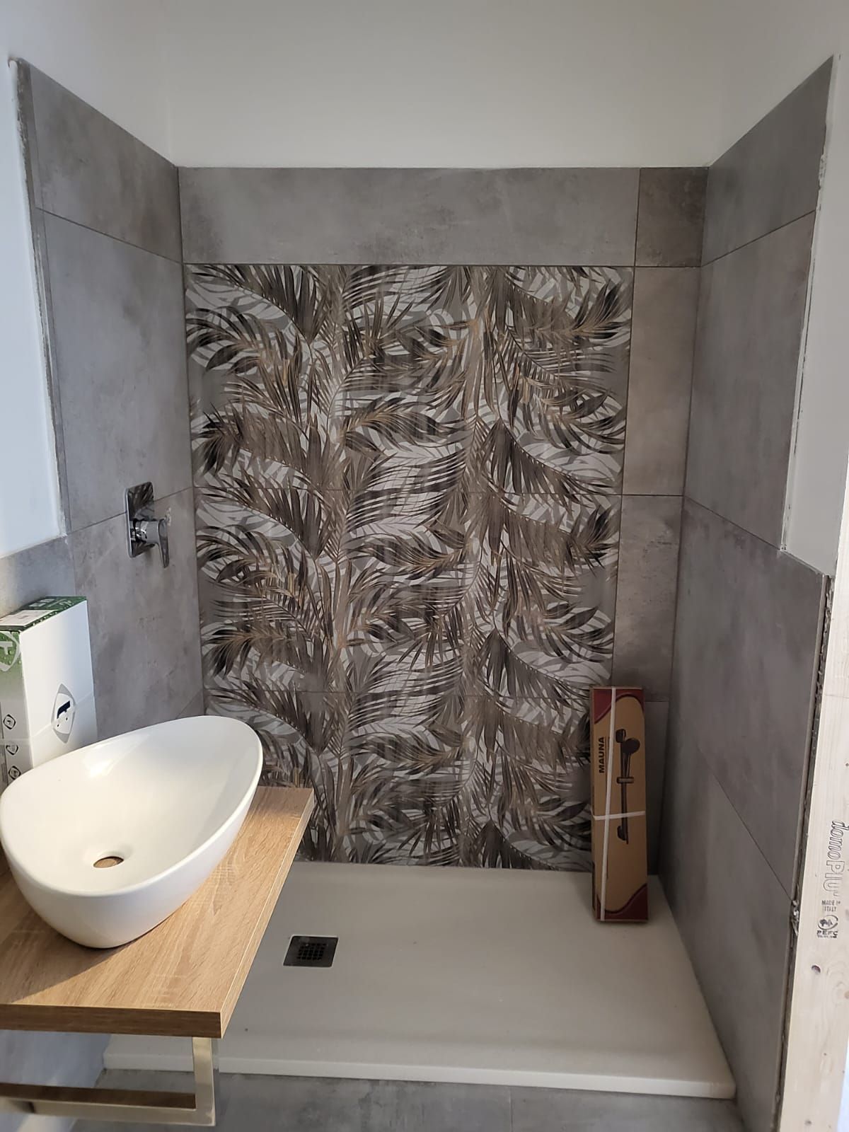 Doccia con parete piastrellata con motivo decorativo a foglie, piastrelle grigie circostanti, lavandino e mensola in legno.