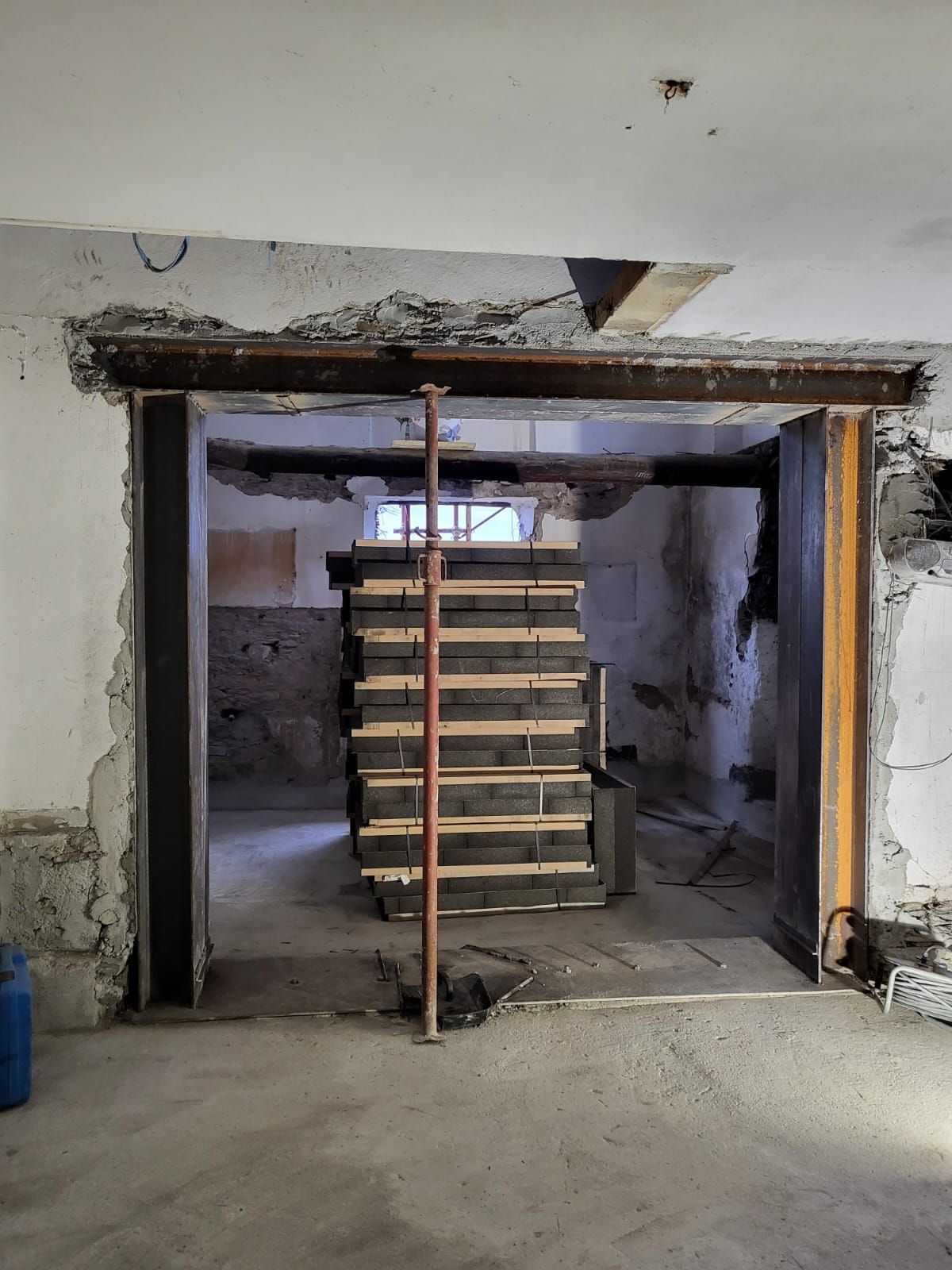 Ristrutturazione di una stanza con travi in acciaio a sostegno di un'apertura. Sono visibili puntelli in legno e una catasta di legna.