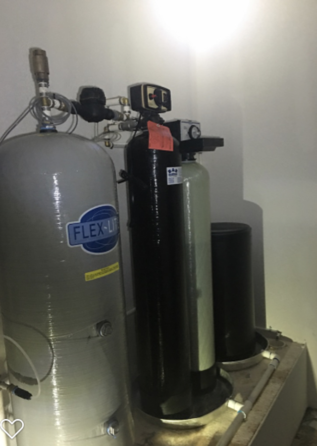 Water softener and filtration tanks in a utility room, including a gray cylinder and black tanks.