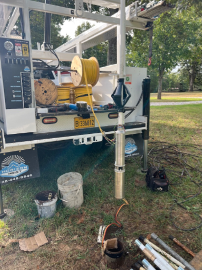 Truck-mounted well drilling rig with yellow equipment and drill pipe on a grassy residential site