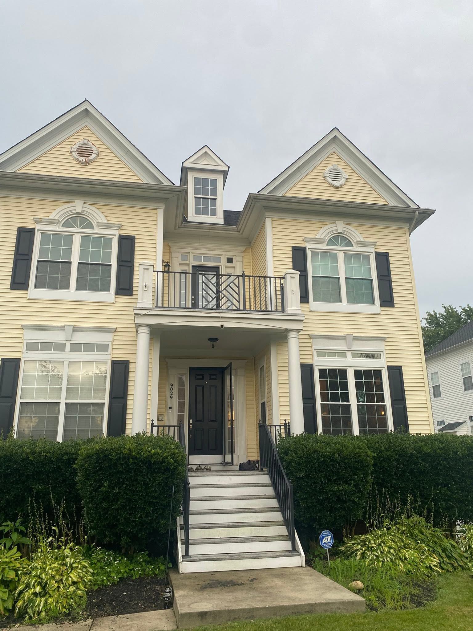 Two-story beige townhouse with black shutters, white trim, and a front staircase behind hedges